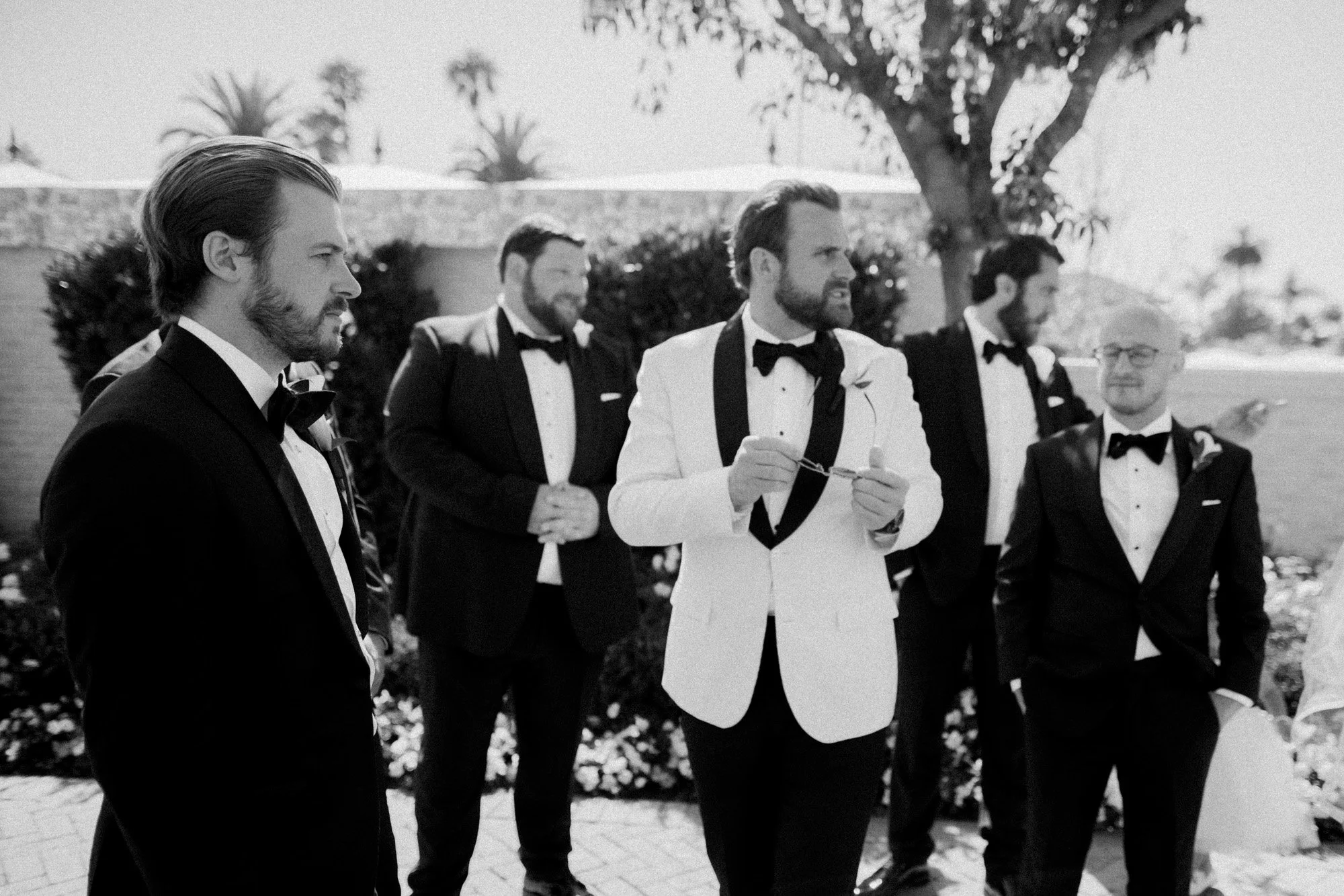 Black and white photo of groom and groomsmen in tuxedos standing outdoors at Rosewood Miramar Beach wedding, photographed by Michael & Anna Costa