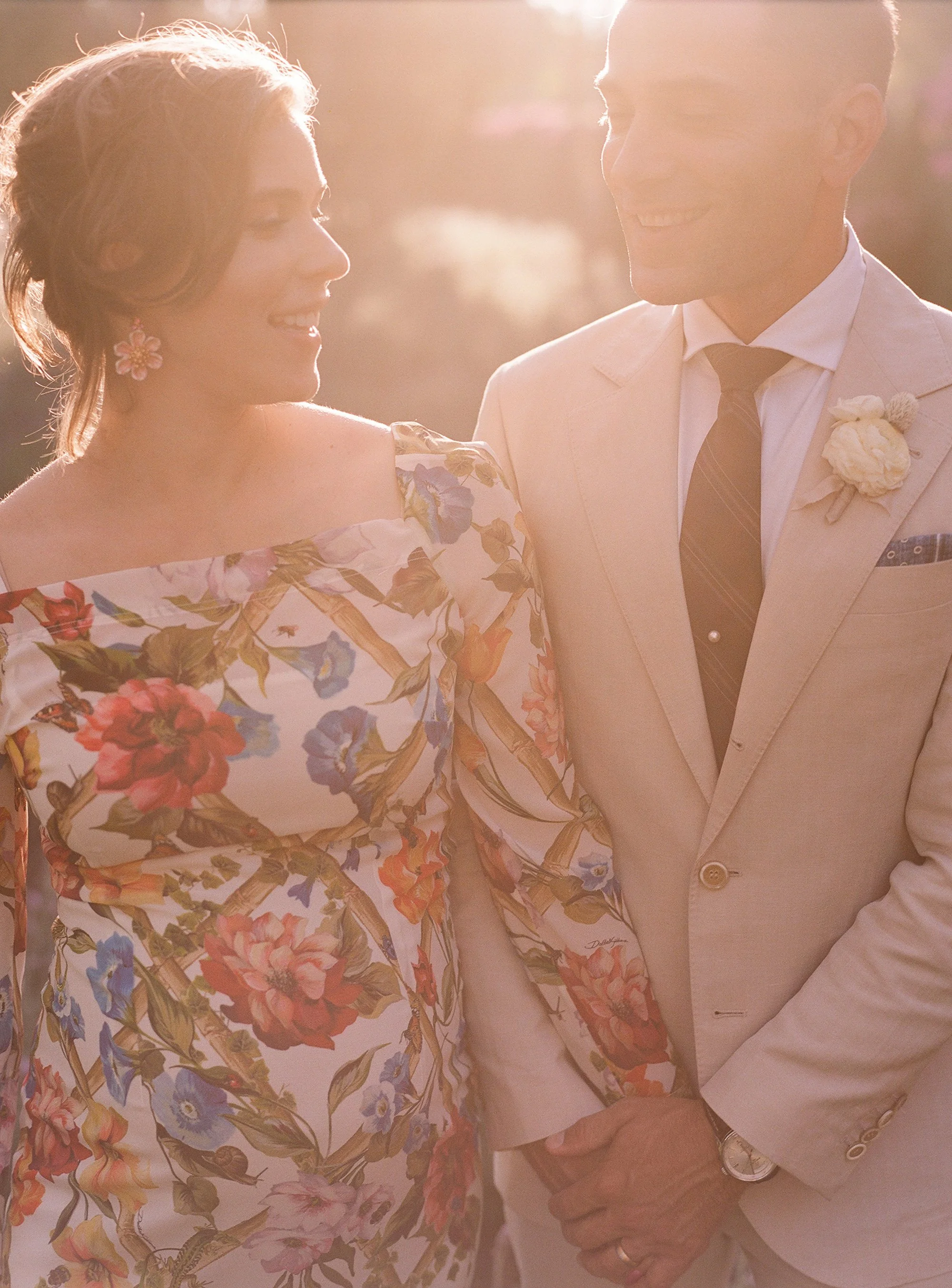 Couple smiling at each other in soft sunlight, bride in a floral dress and groom in a light suit.