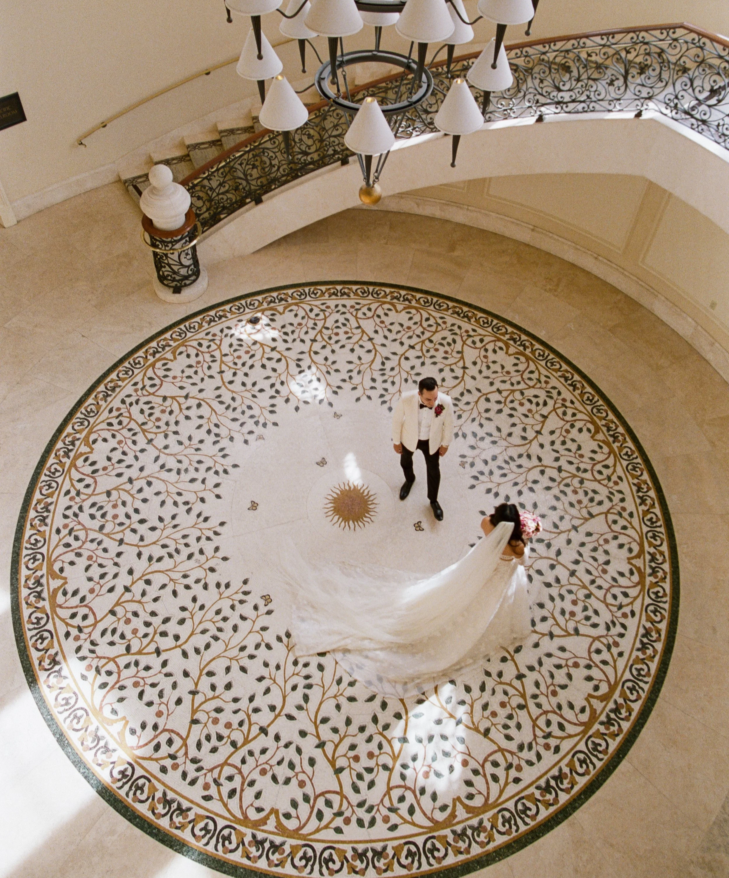 Interior view of a grand staircase and a circular patterned marble floor, featuring an elegant chandelier hanging from above.