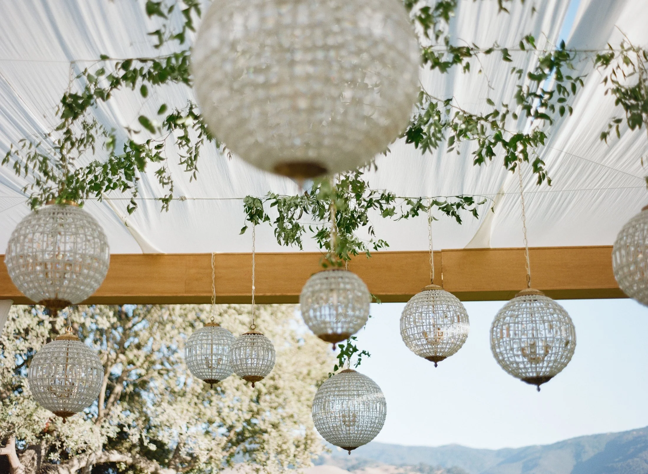 Hanging glass lanterns and greenery decor under a pergola at an outdoor wedding.