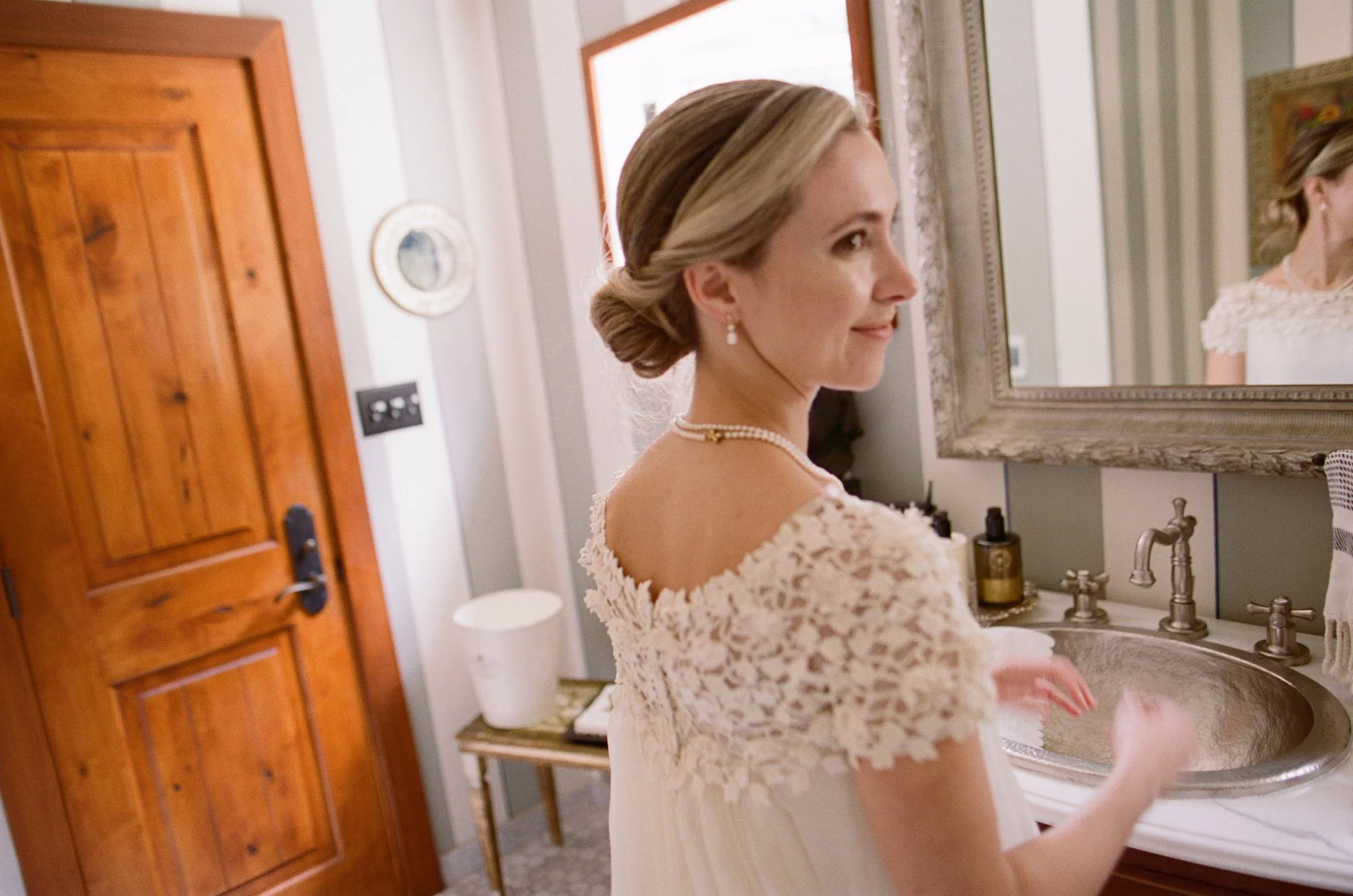 Bride with a low bun hairstyle and lace dress, smiling softly in a sunlit room.