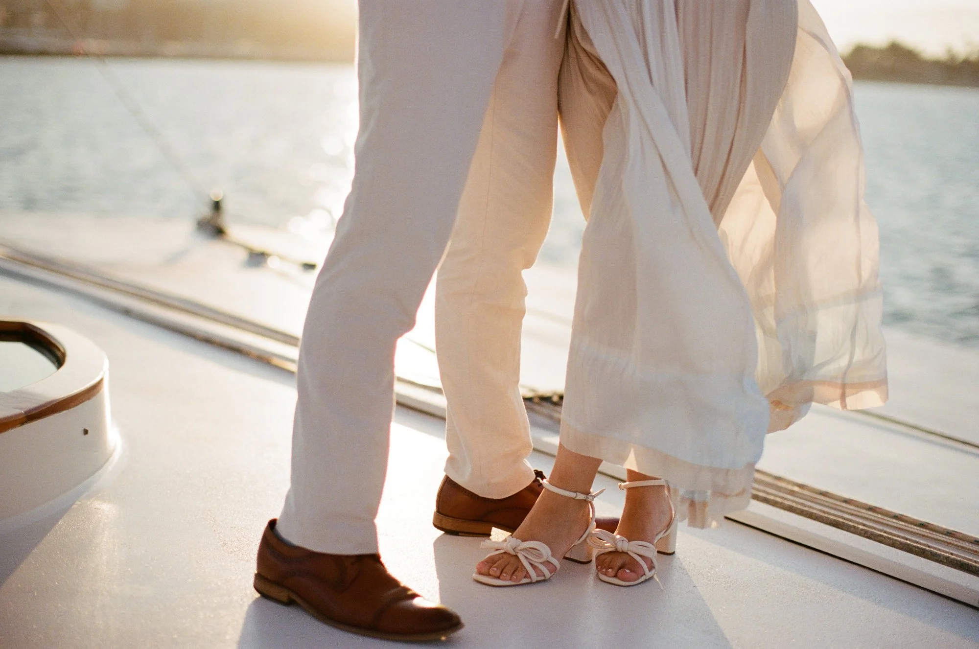 Close-up of couple’s feet on the yacht deck, bride’s dress flowing in the wind.