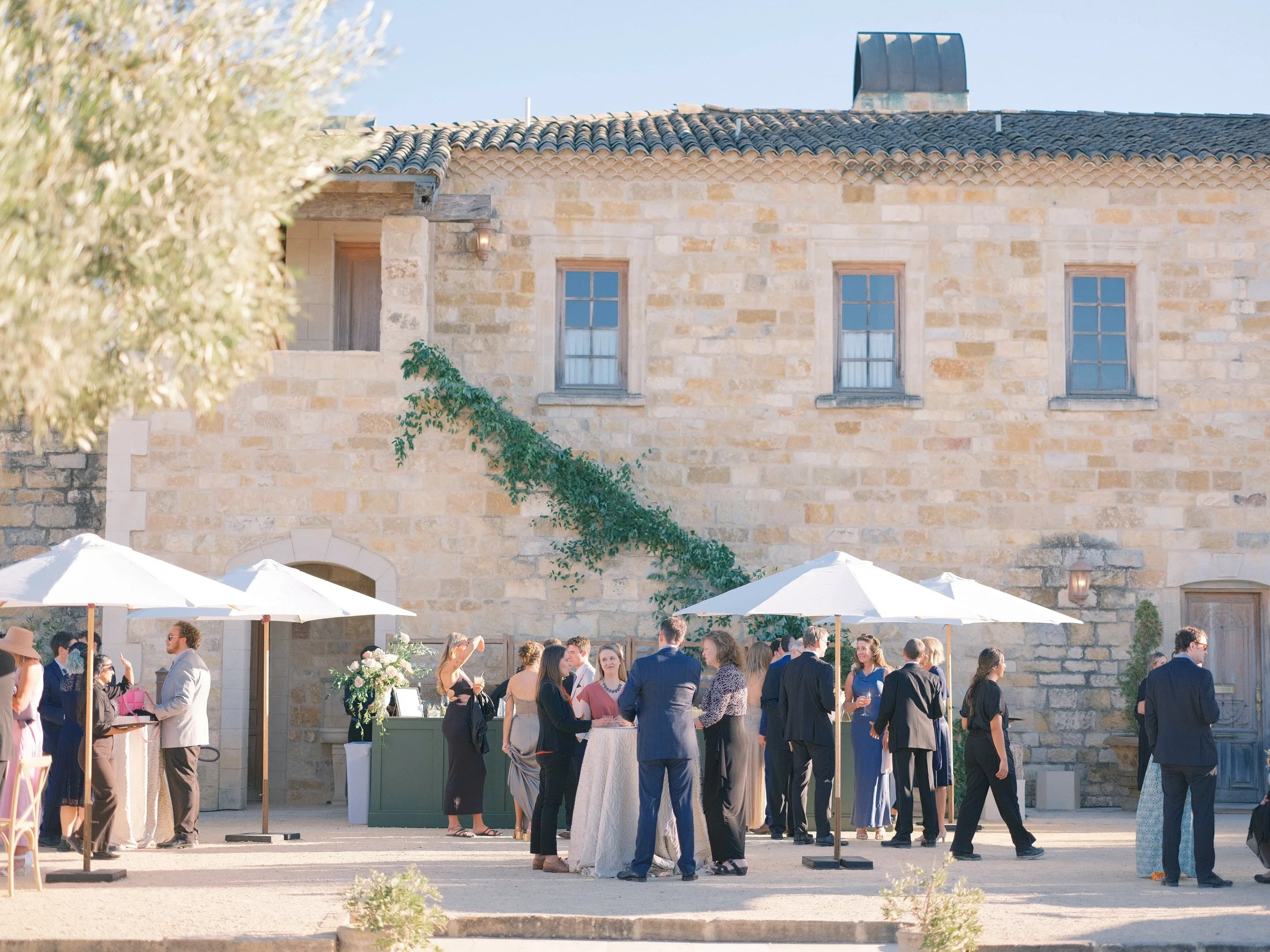 Wedding guests gathered outside a rustic stone building with umbrellas.