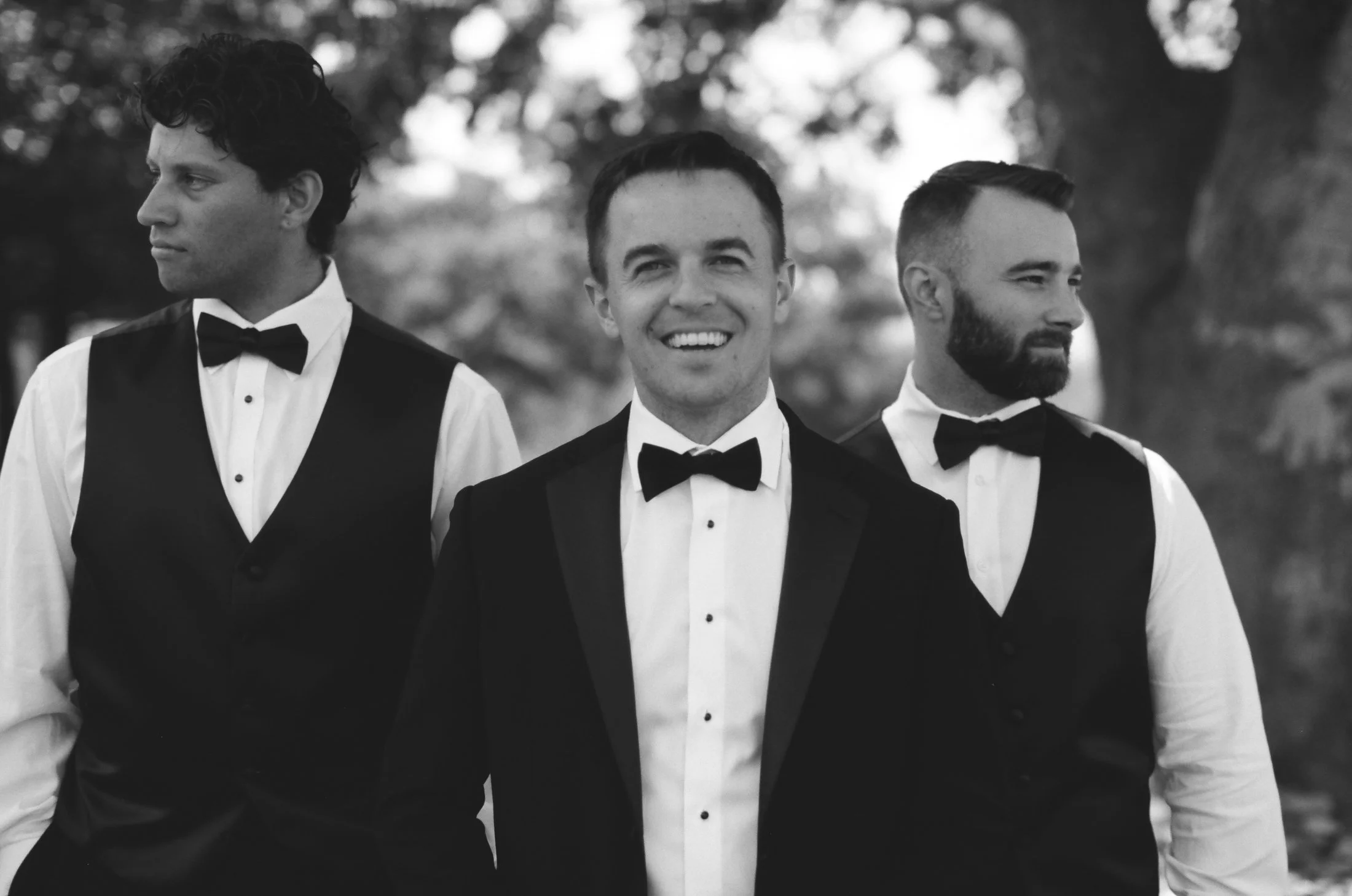 Black and white photo of groom and groomsmen in tuxedos smiling outdoors at Sunstone Villa wedding, photographed by Michael & Anna Costa