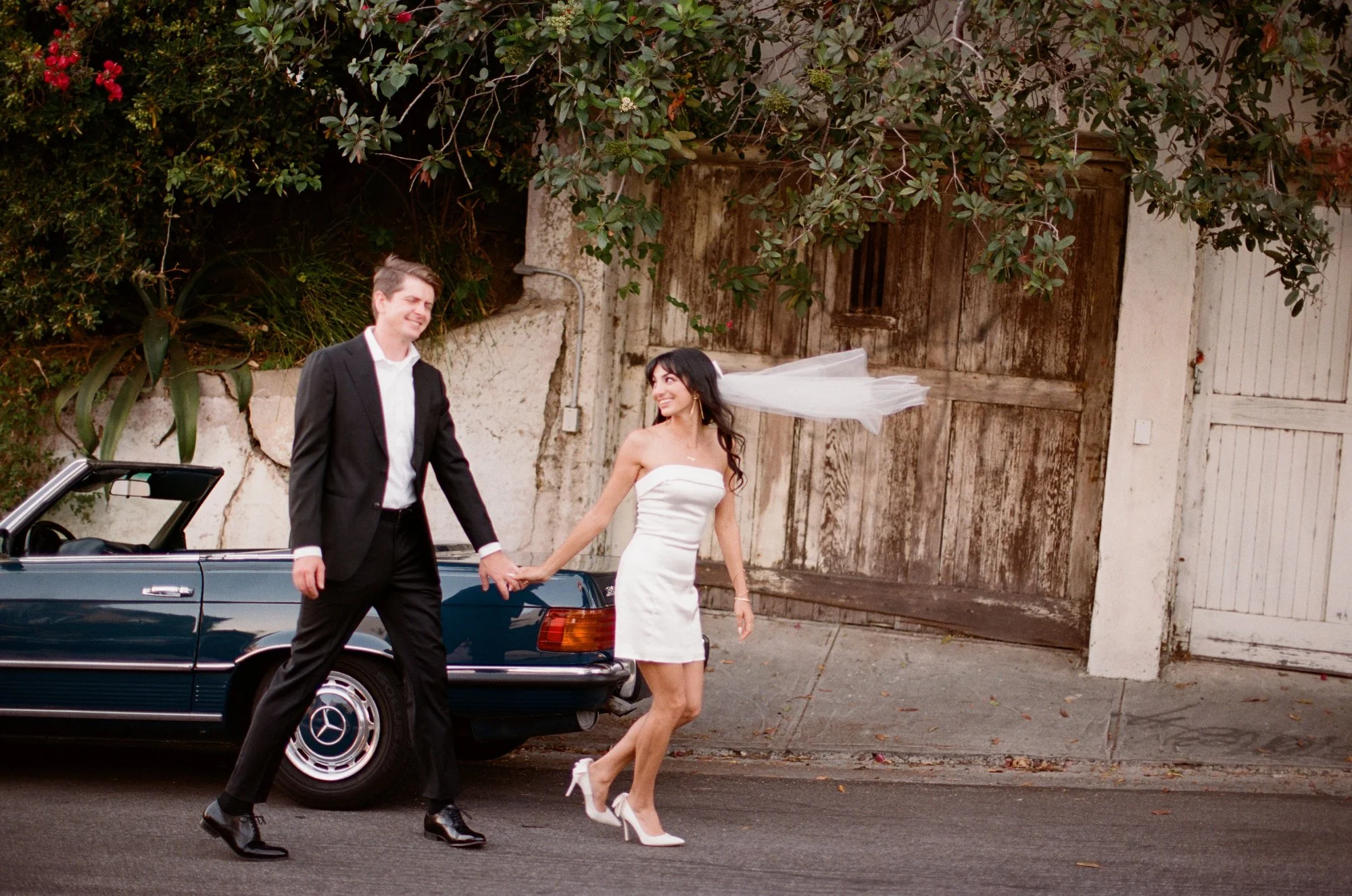Couple sitting on a vintage car, bride in a white dress and groom in a tuxedo, with greenery in the background.