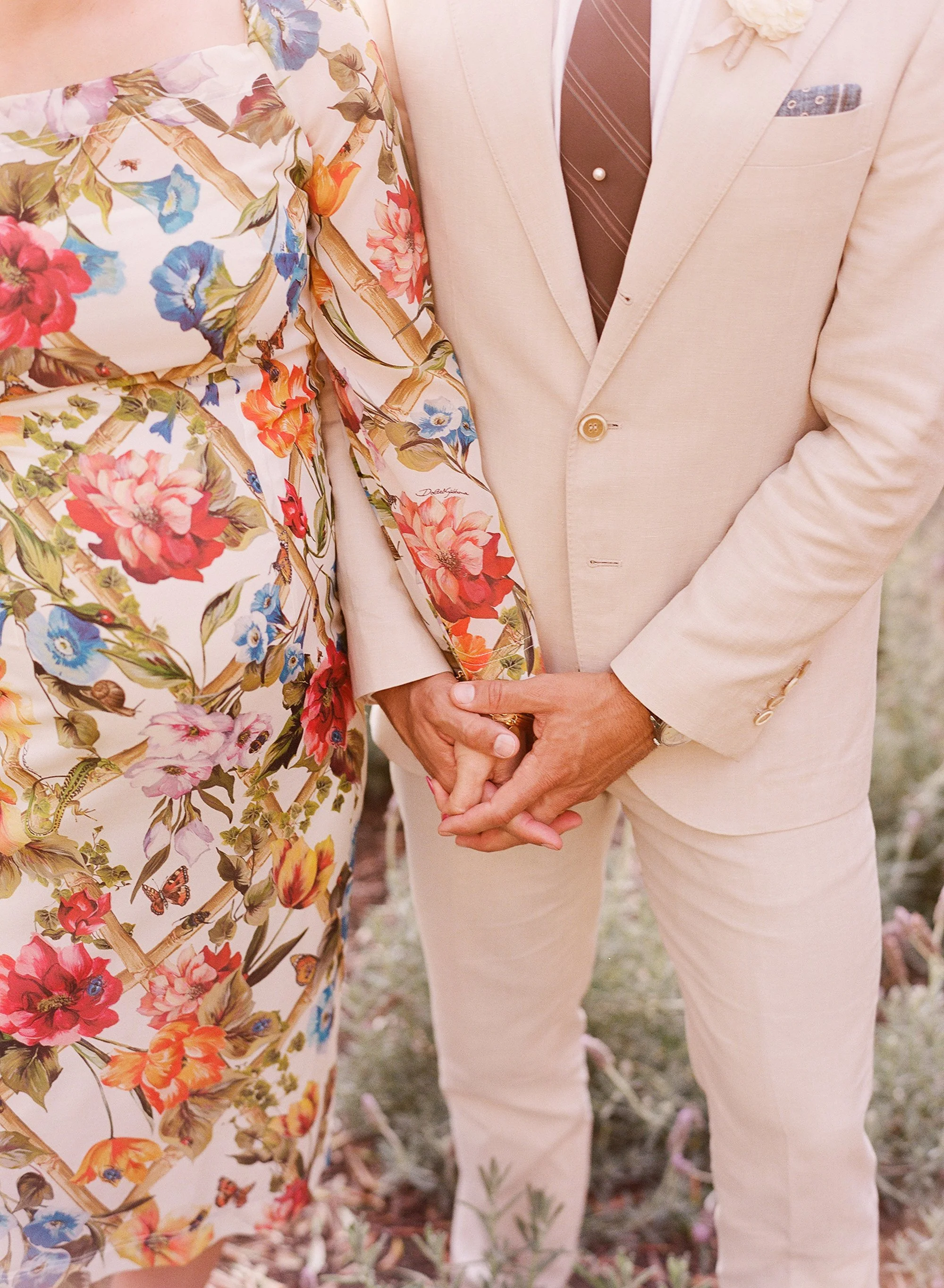 Close-up of couple holding hands, bride in a floral dress and groom in a beige suit.