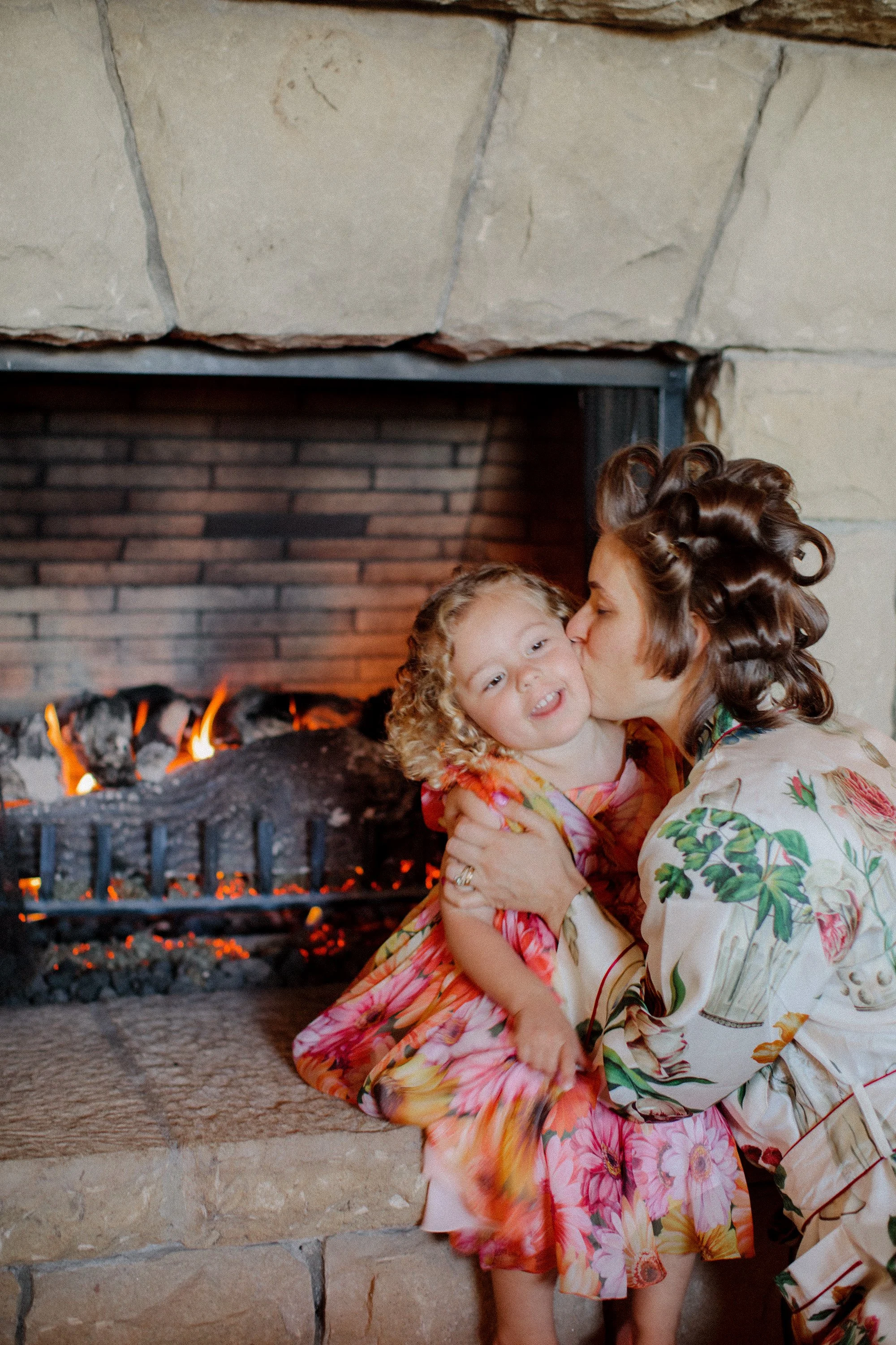 Mother and daughter in floral dresses cuddling by a fireplace.