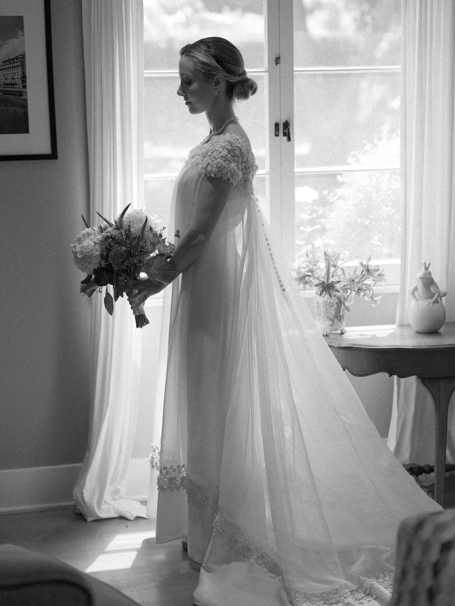 Black and white photo of bride in a lace gown holding a bouquet by a window.