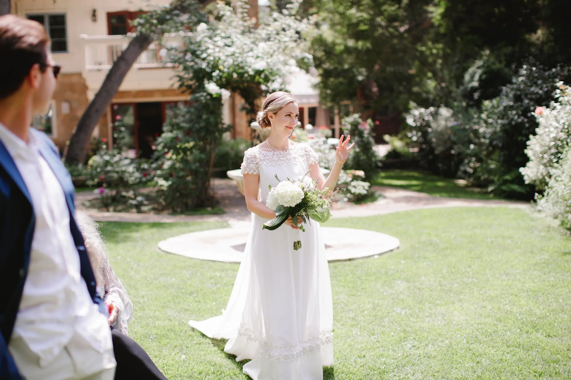 Bride standing outdoors in sunlight, holding a bouquet and smiling.