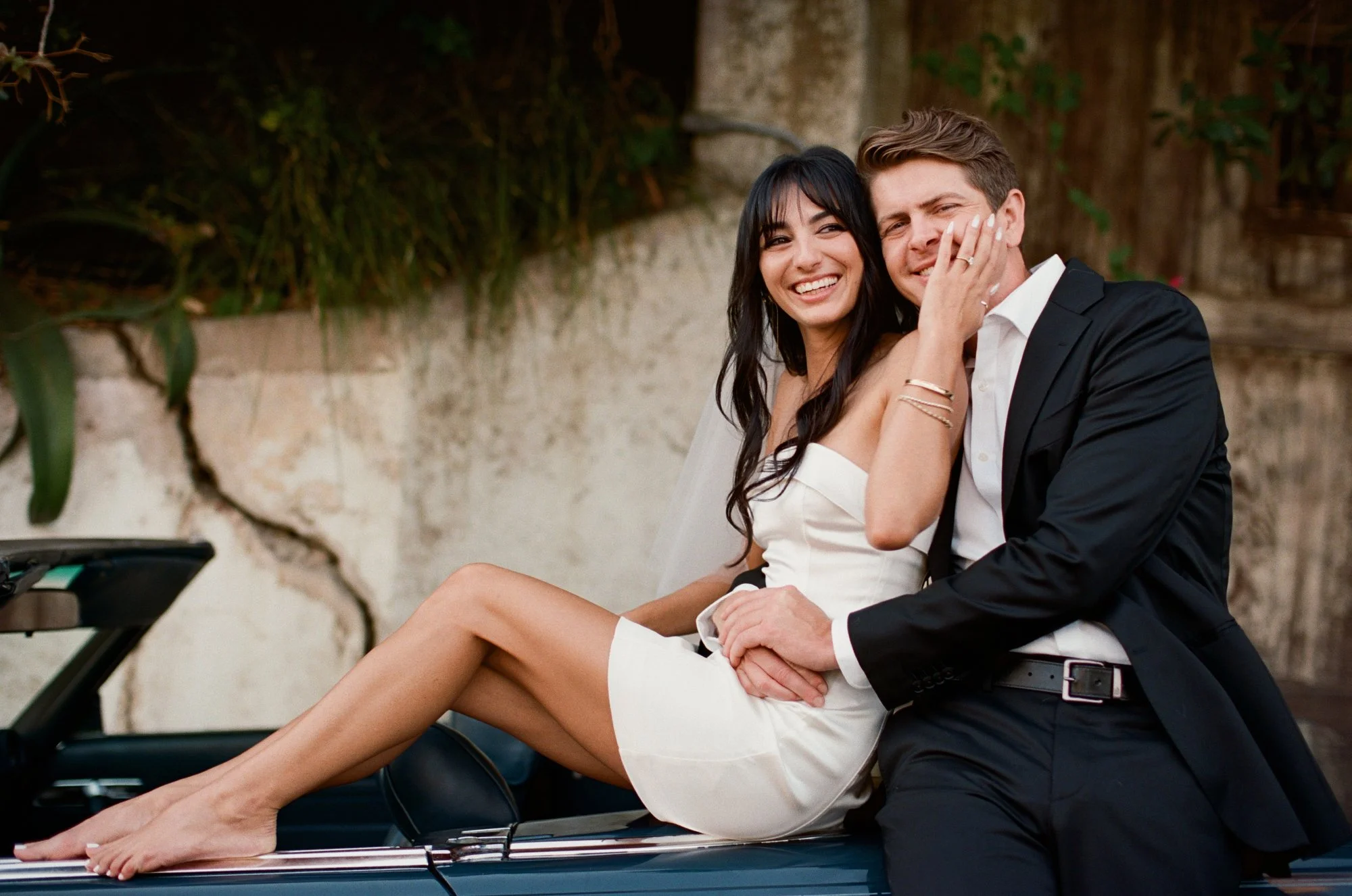 Couple sitting together on a blue vintage convertible car, bride in a white dress and groom in a black tuxedo.