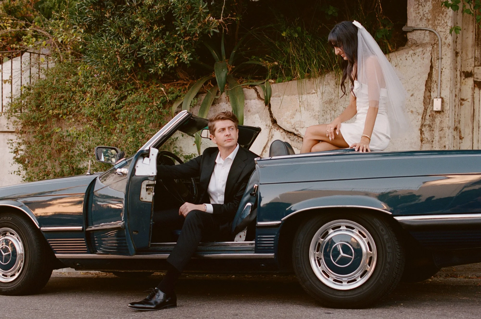 Couple standing by a vintage car, groom in a tuxedo and bride in a white dress.