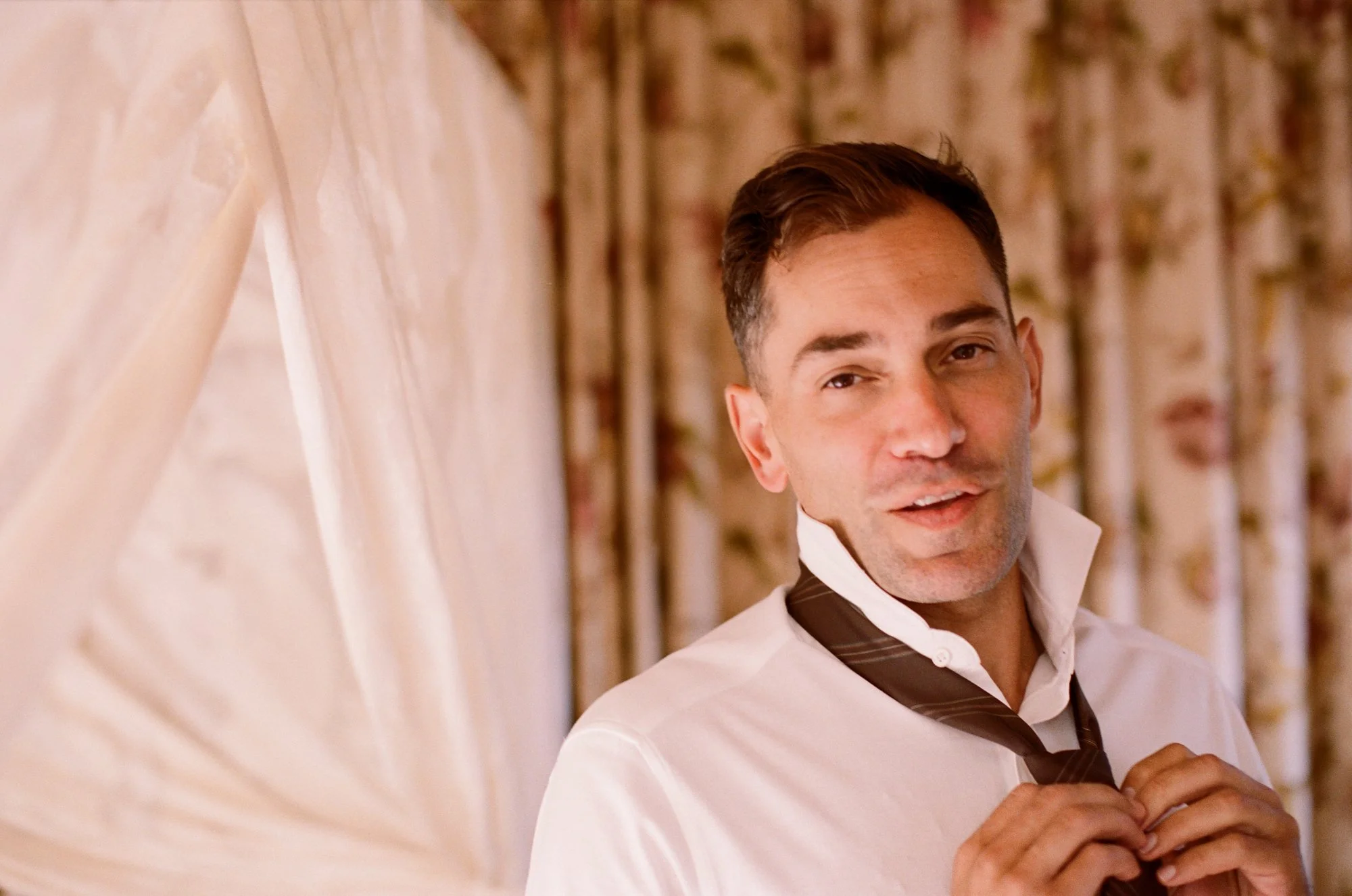 Man in a white shirt putting on a tie, standing indoors by a curtain.