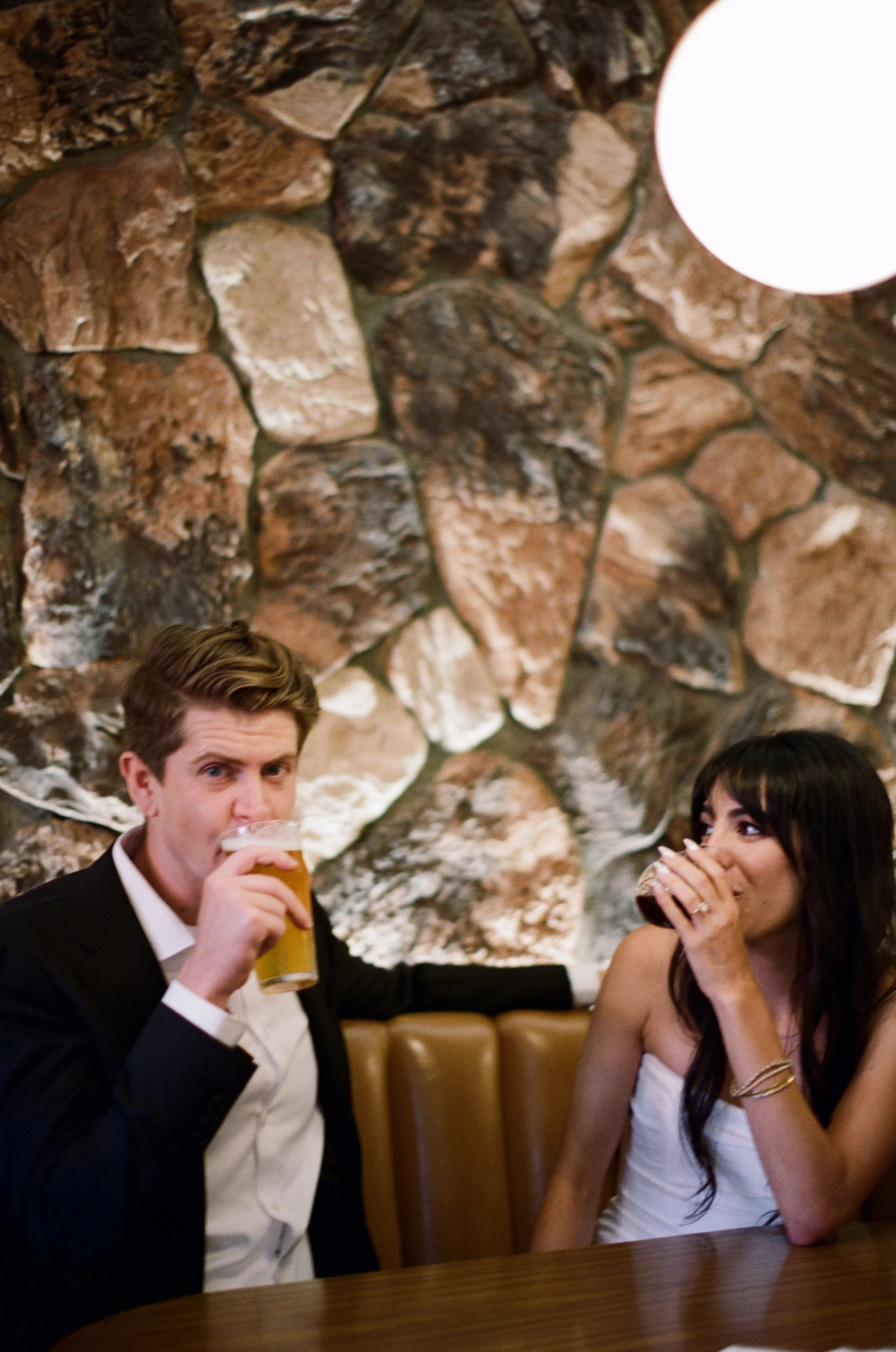 Couple sitting close together, bride in a white dress and groom in a tuxedo, against a stone wall.