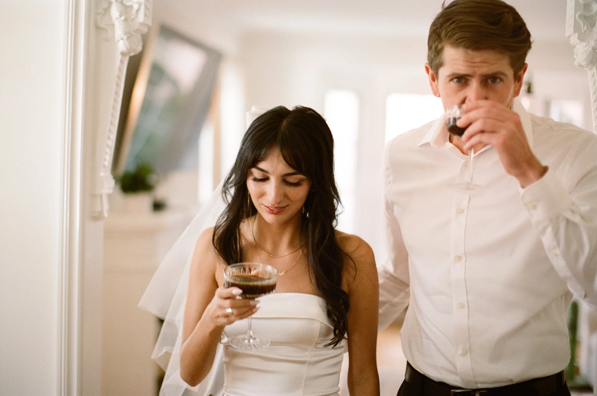 Couple indoors, the woman in a white dress smiling while holding a glass of wine, and the man in a white shirt standing beside her in a softly lit room.