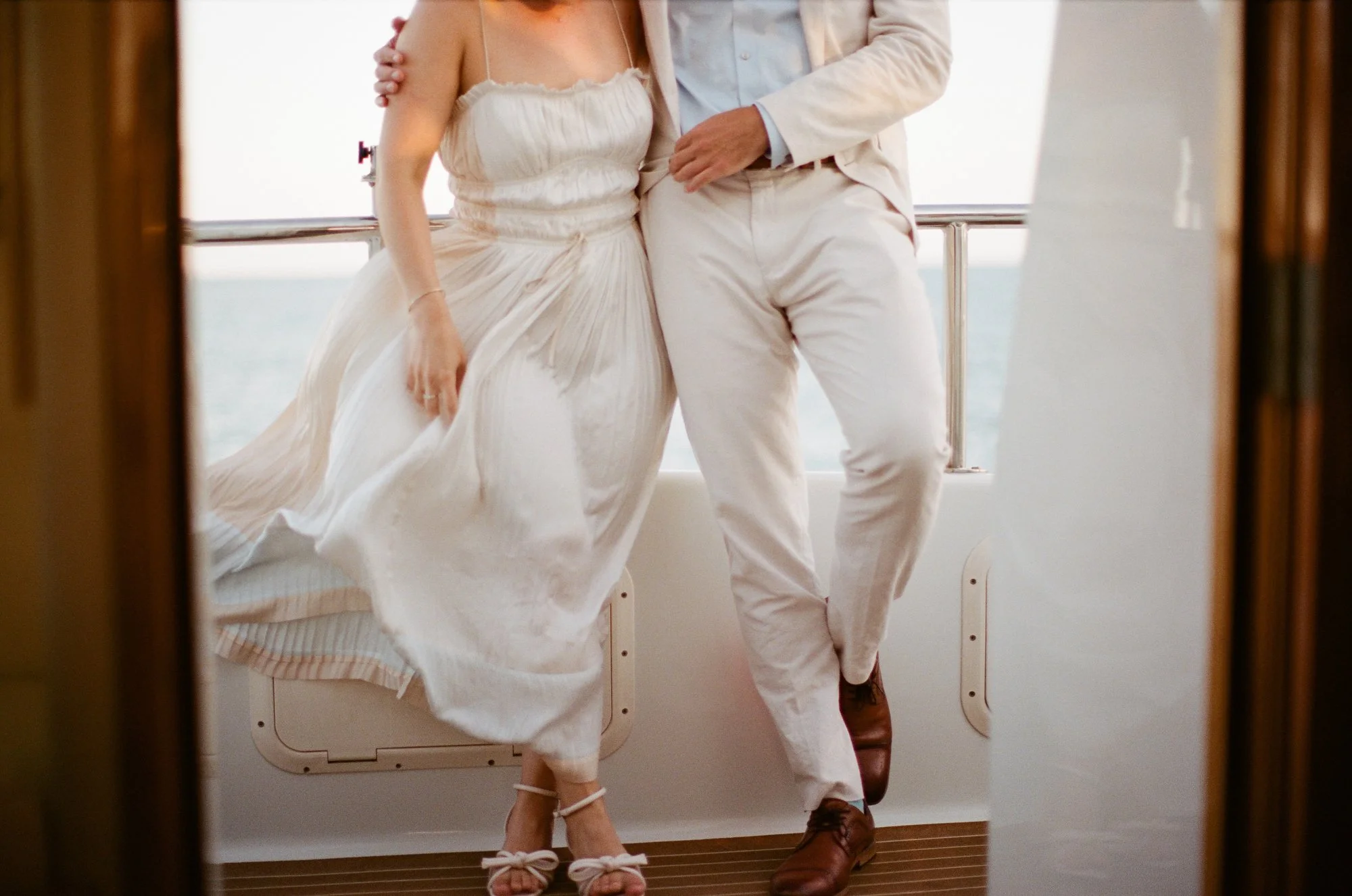 Bride and groom walking hand in hand on a yacht deck, both dressed in white, with the ocean in the background.