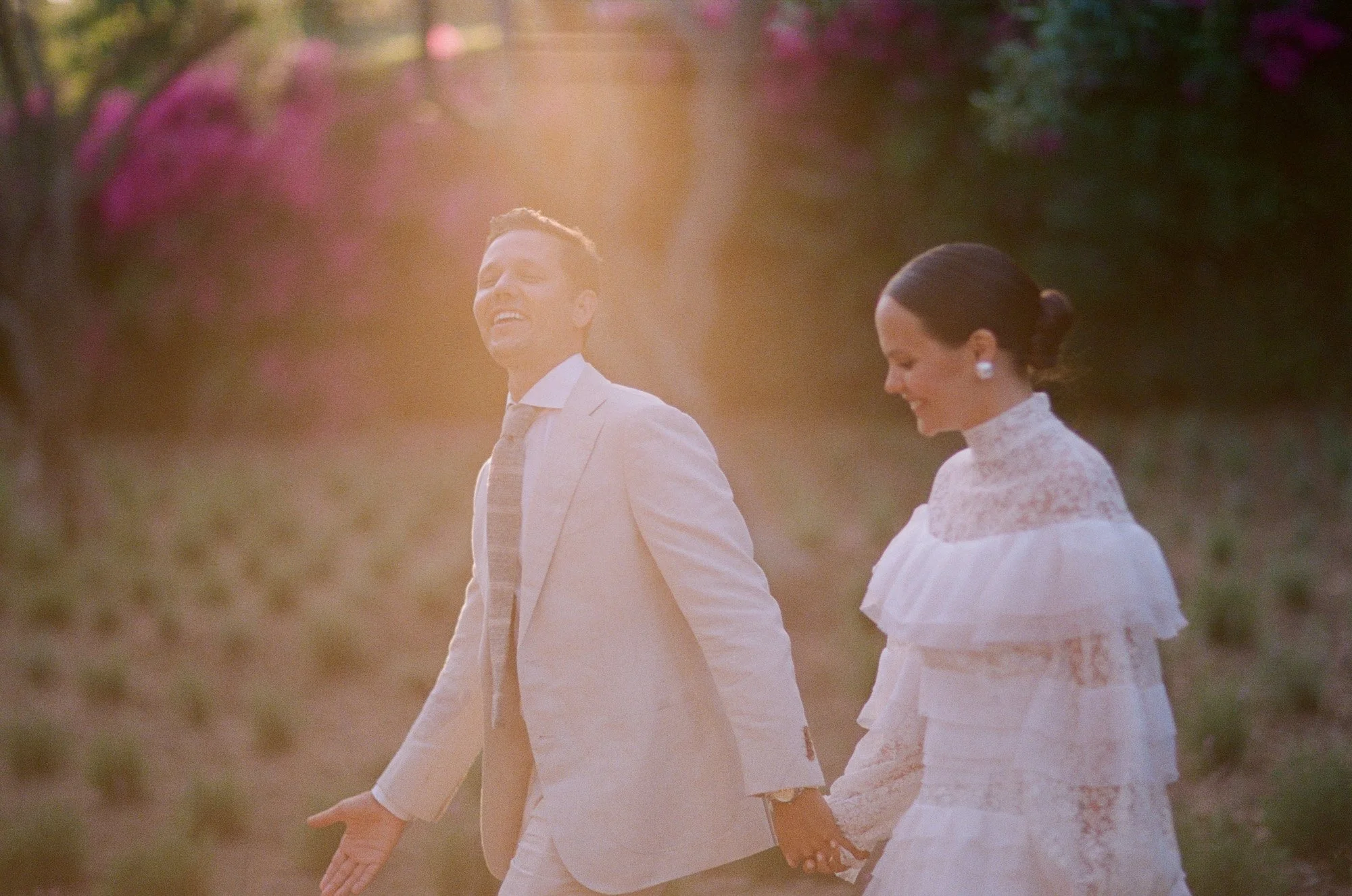 Bride and groom walking hand in hand in sunlight with bougainvillea in the background at San Ysidro Ranch, photographed by Michael & Anna Costa