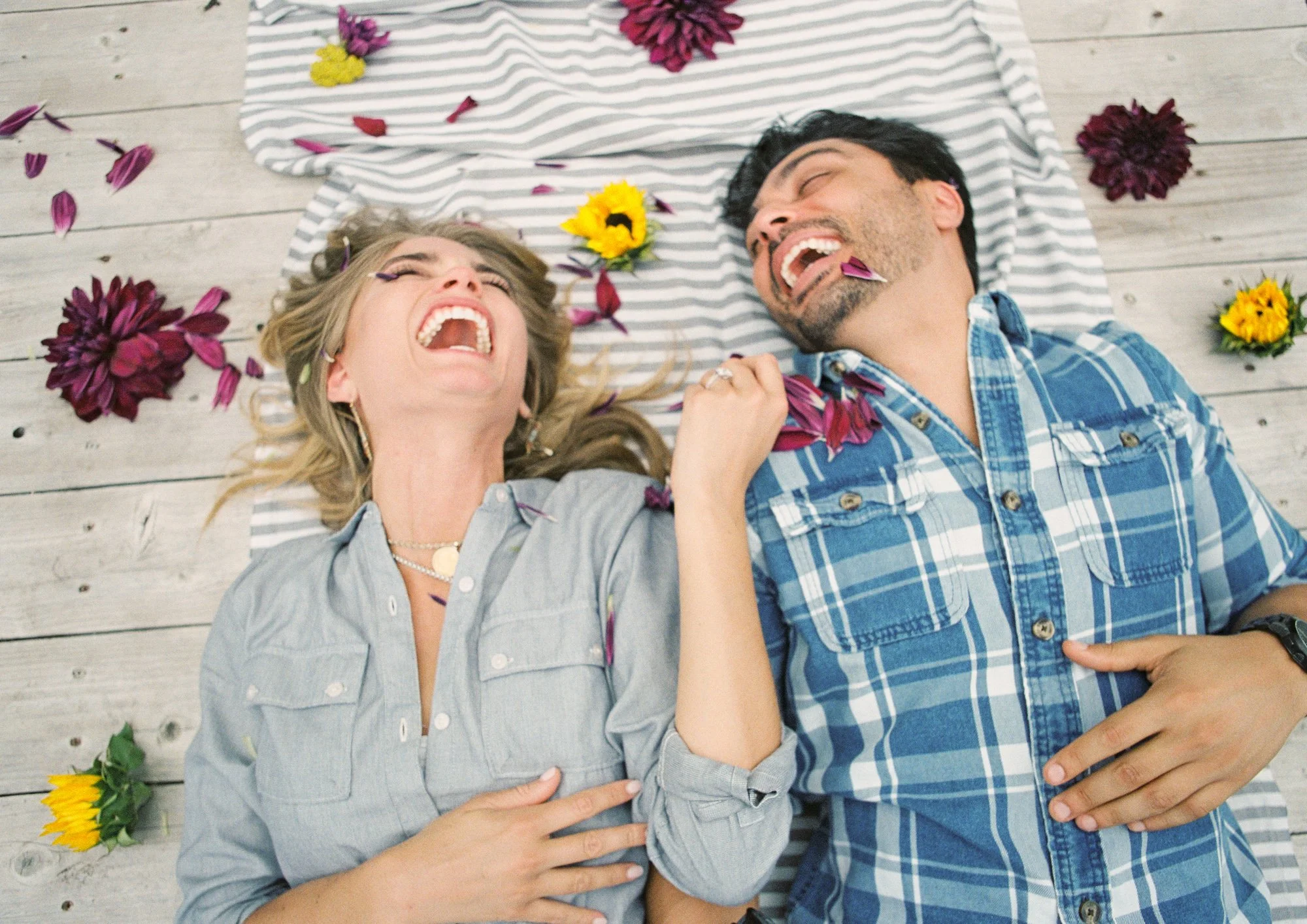 Couple lying on a blanket outdoors, laughing together, surrounded by flowers.