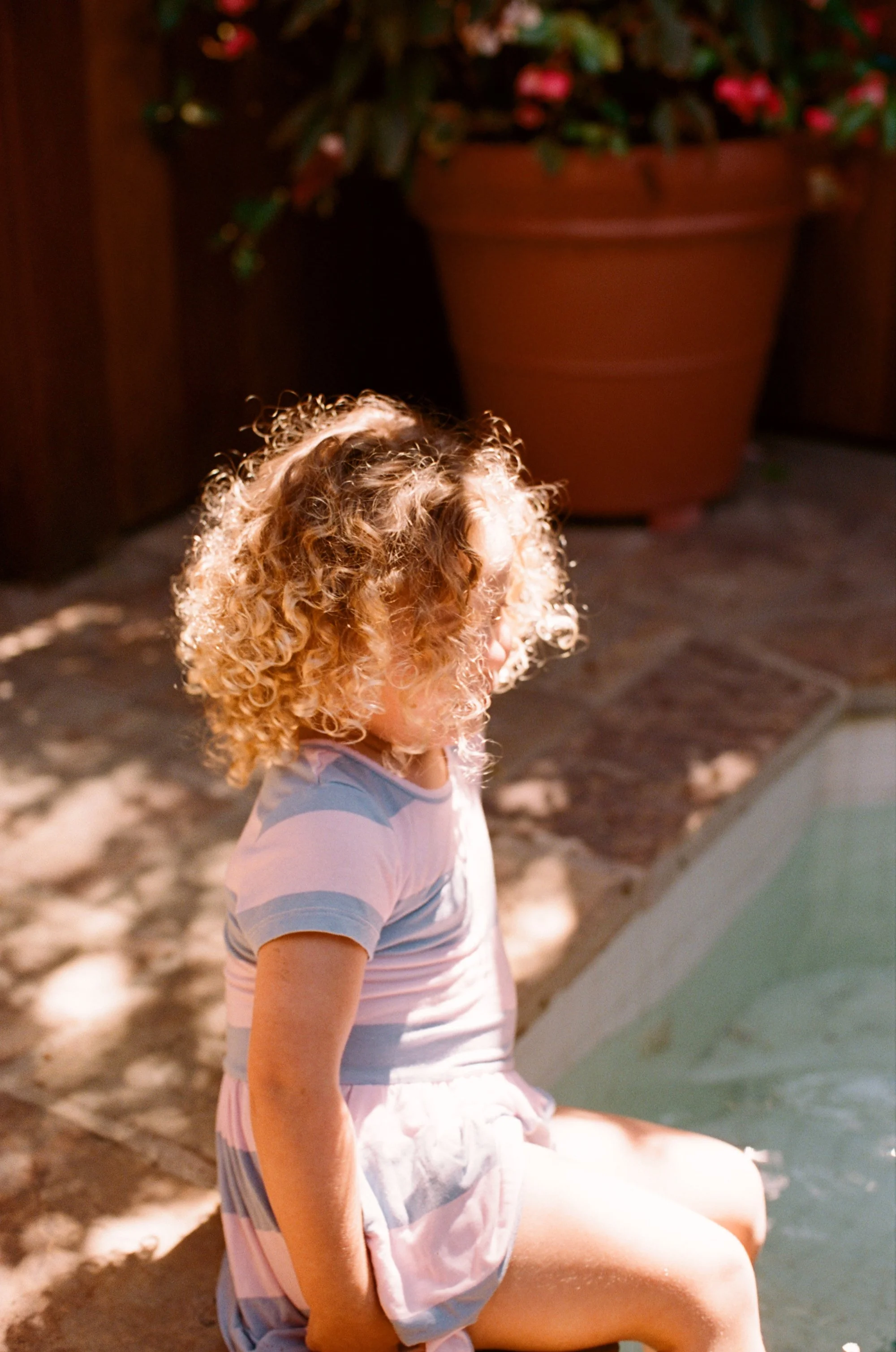 Young child with curly hair wearing a striped shirt, sitting outdoors in sunlight.