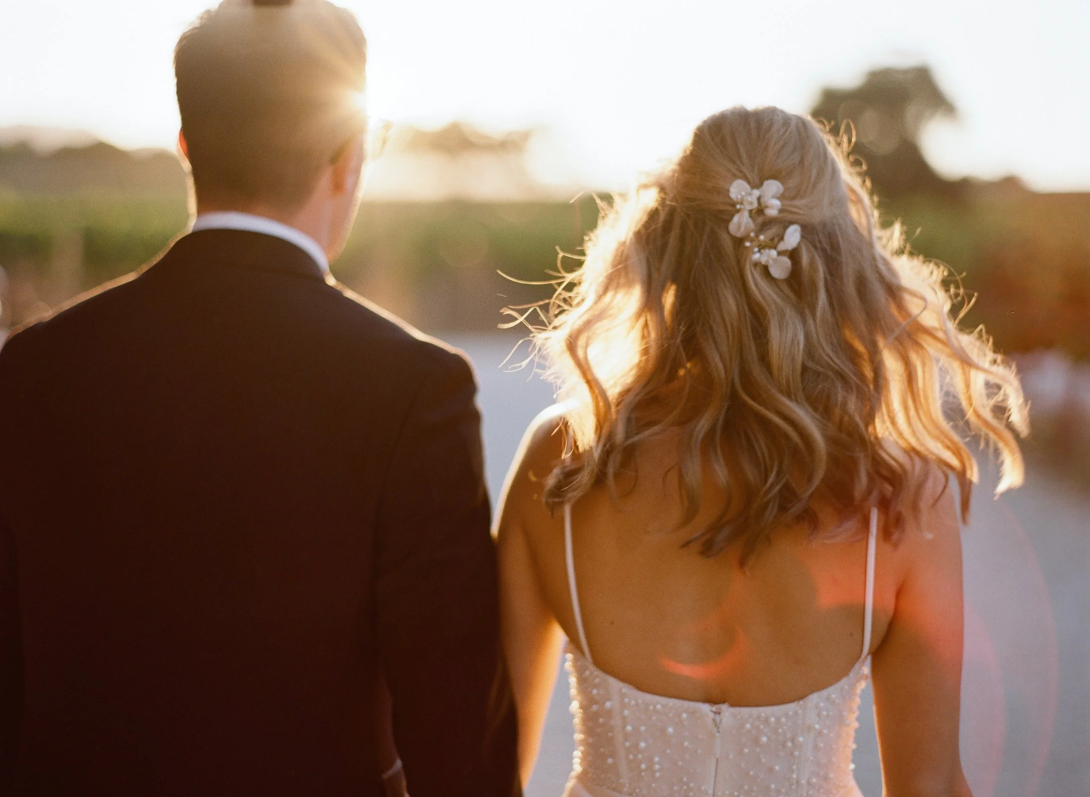Bride and groom from behind, bride’s hair styled with floral accents, golden hour light.