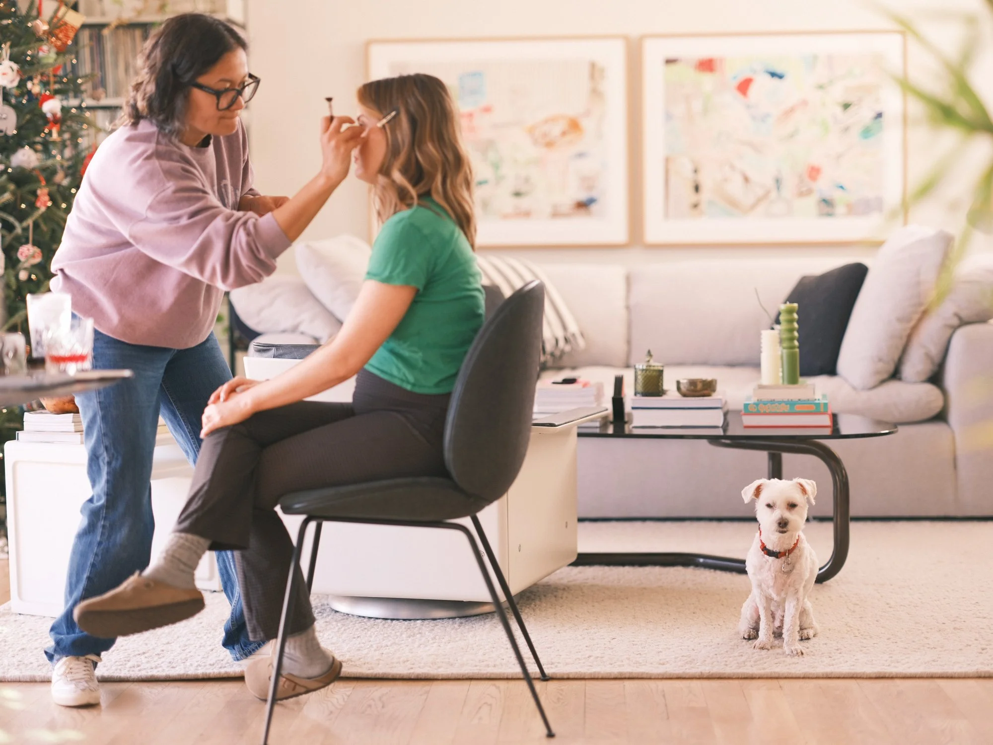 Woman applying makeup to another woman seated in a modern living room, with a small white dog nearby.