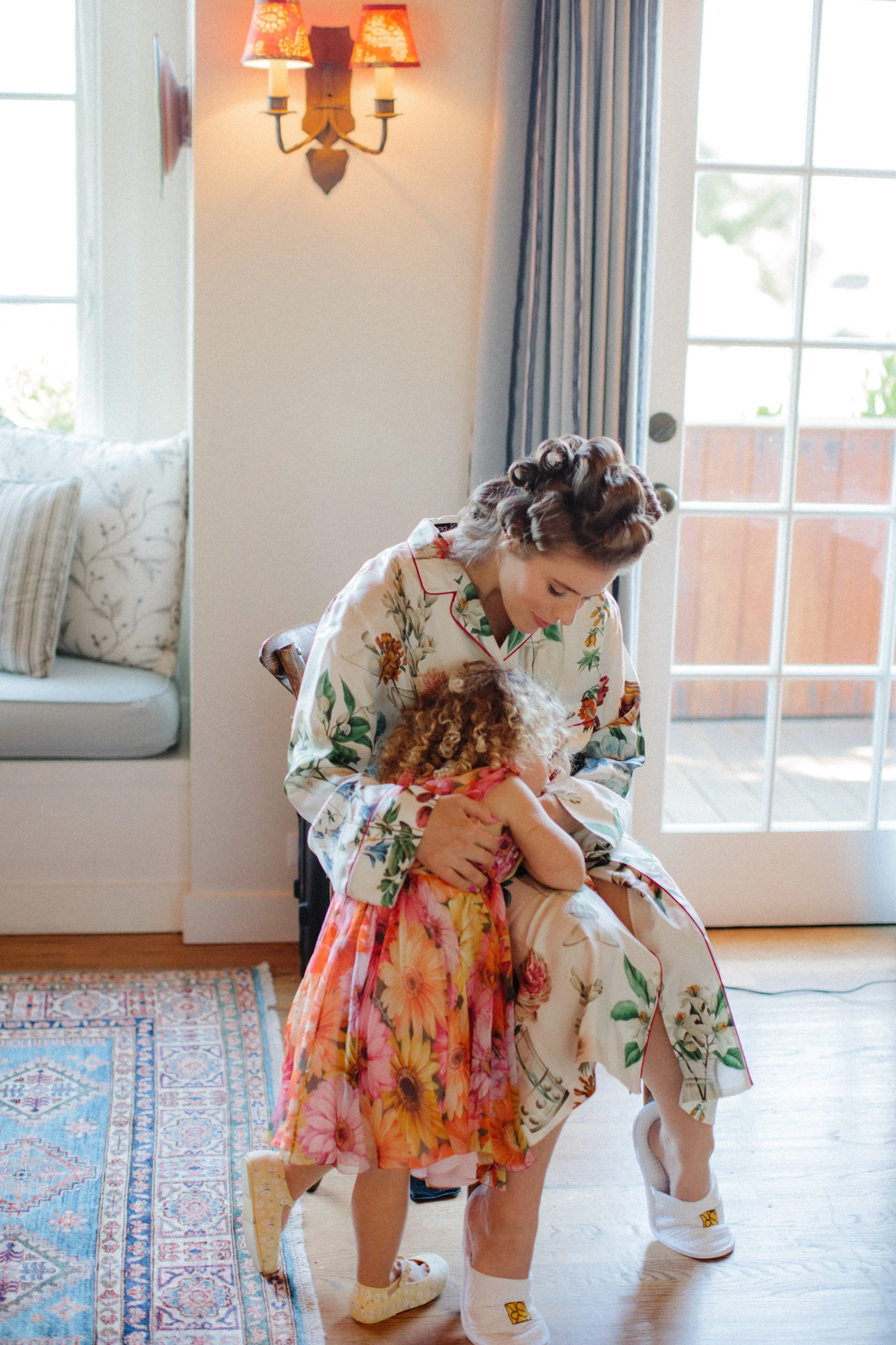 Mother in a floral robe hugging her daughter in a sunlit living room.