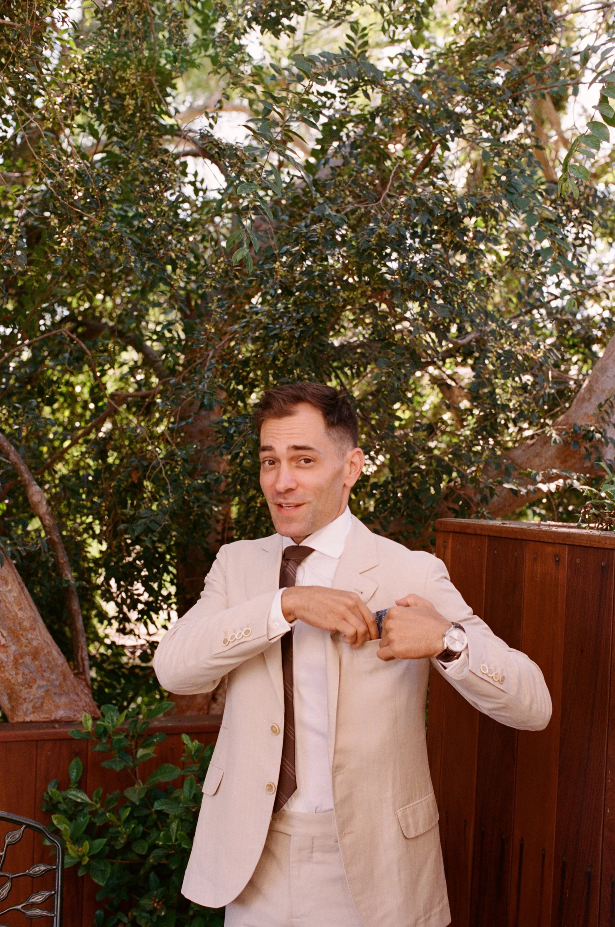 Man in a white shirt and tie standing outdoors, surrounded by trees.