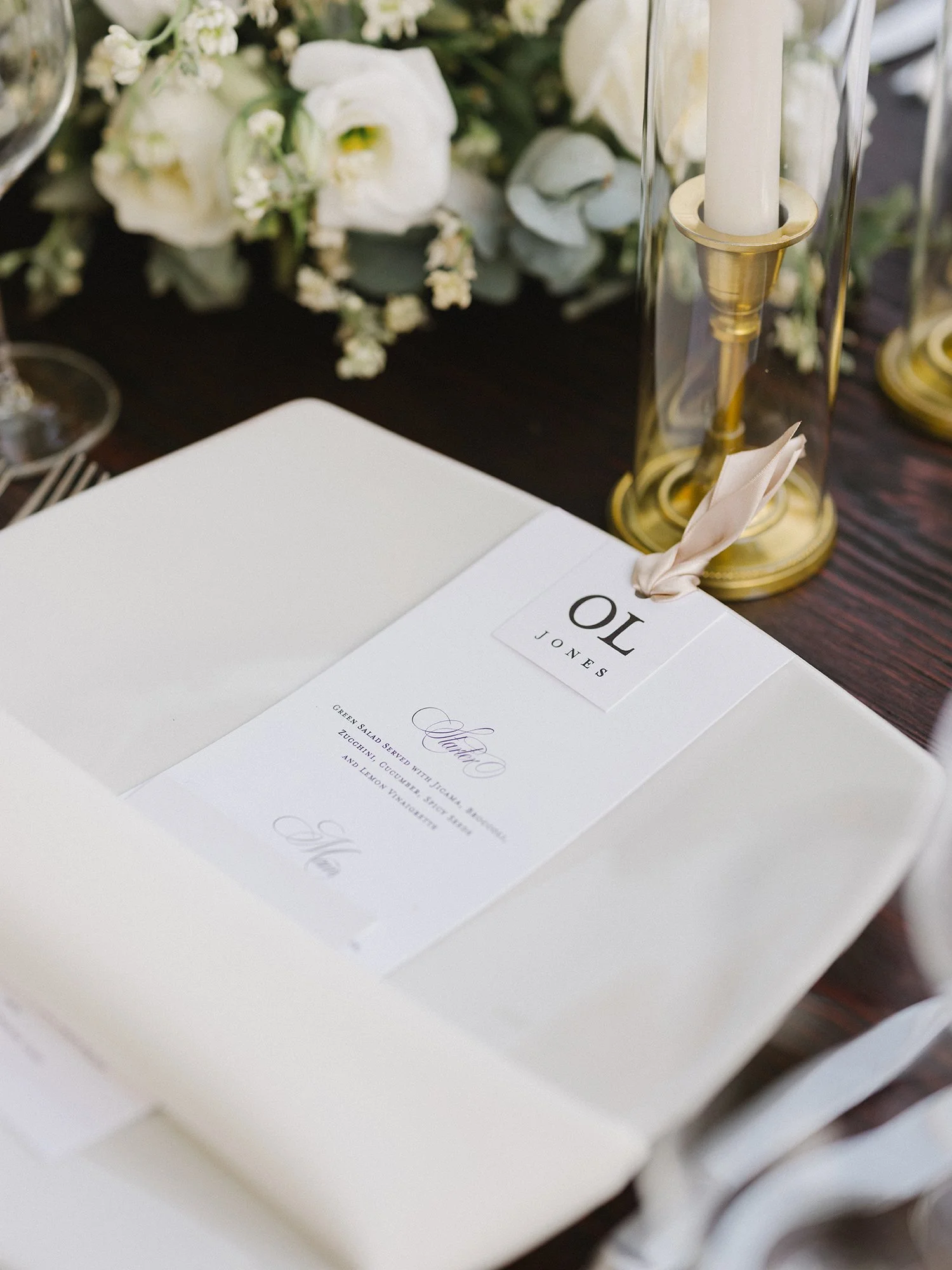 Close-up of elegant wedding table setting with white napkin, personalized menu card, and white floral centerpiece.