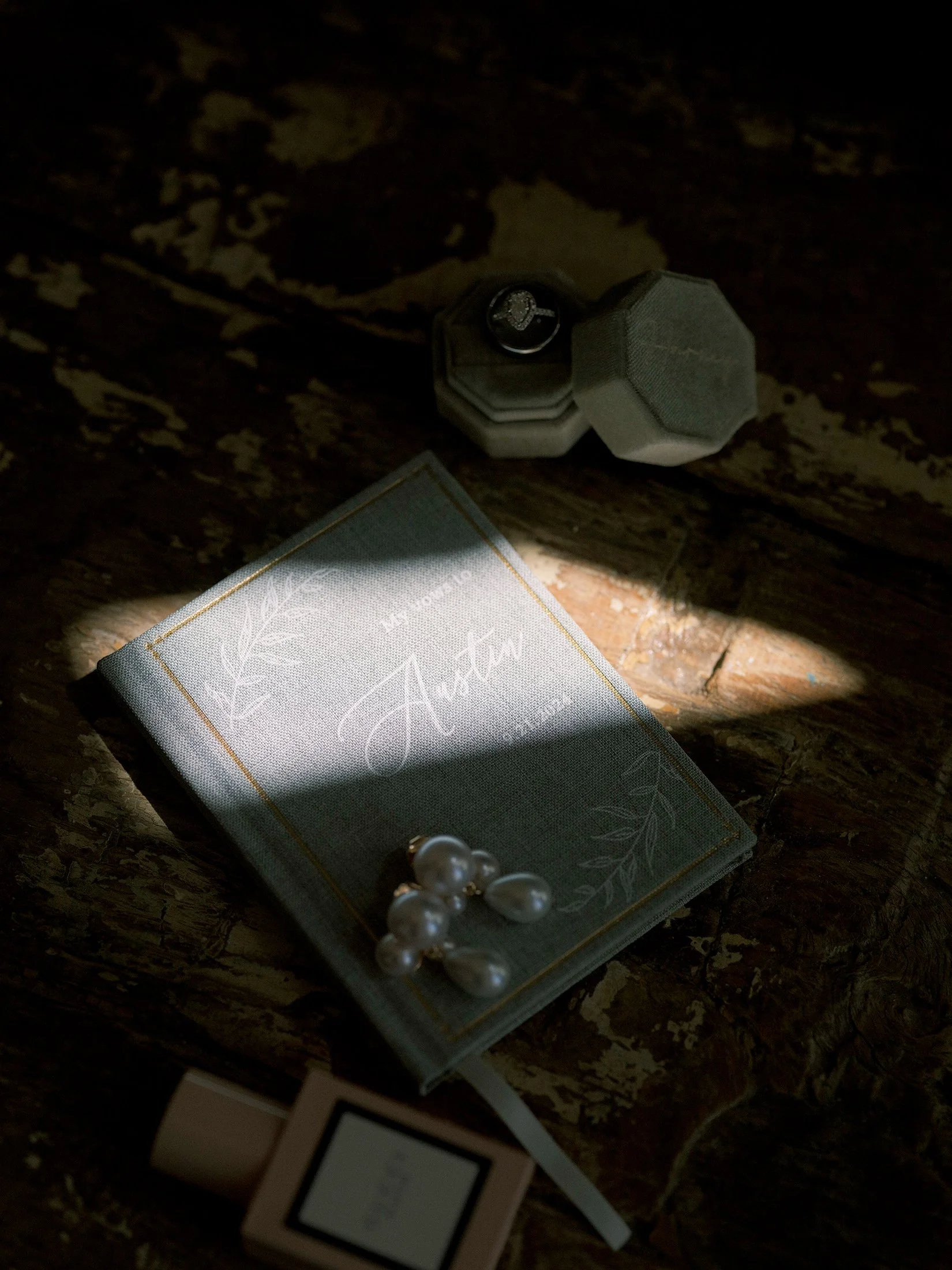 Artistic close-up of a wedding invitation, pearl earrings, and ring box in dramatic light.
