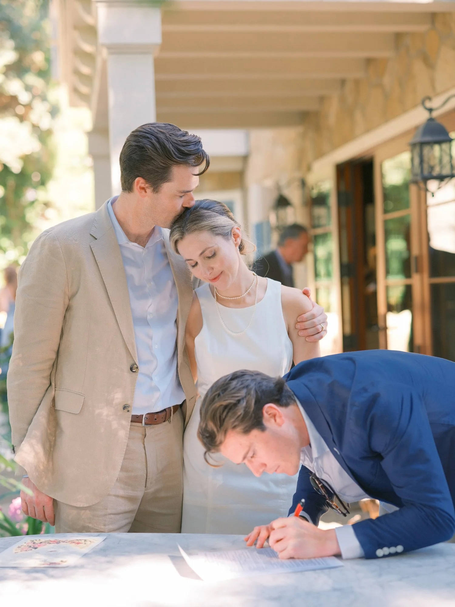 Groom in a tan suit kneeling to greet a young boy outdoors.