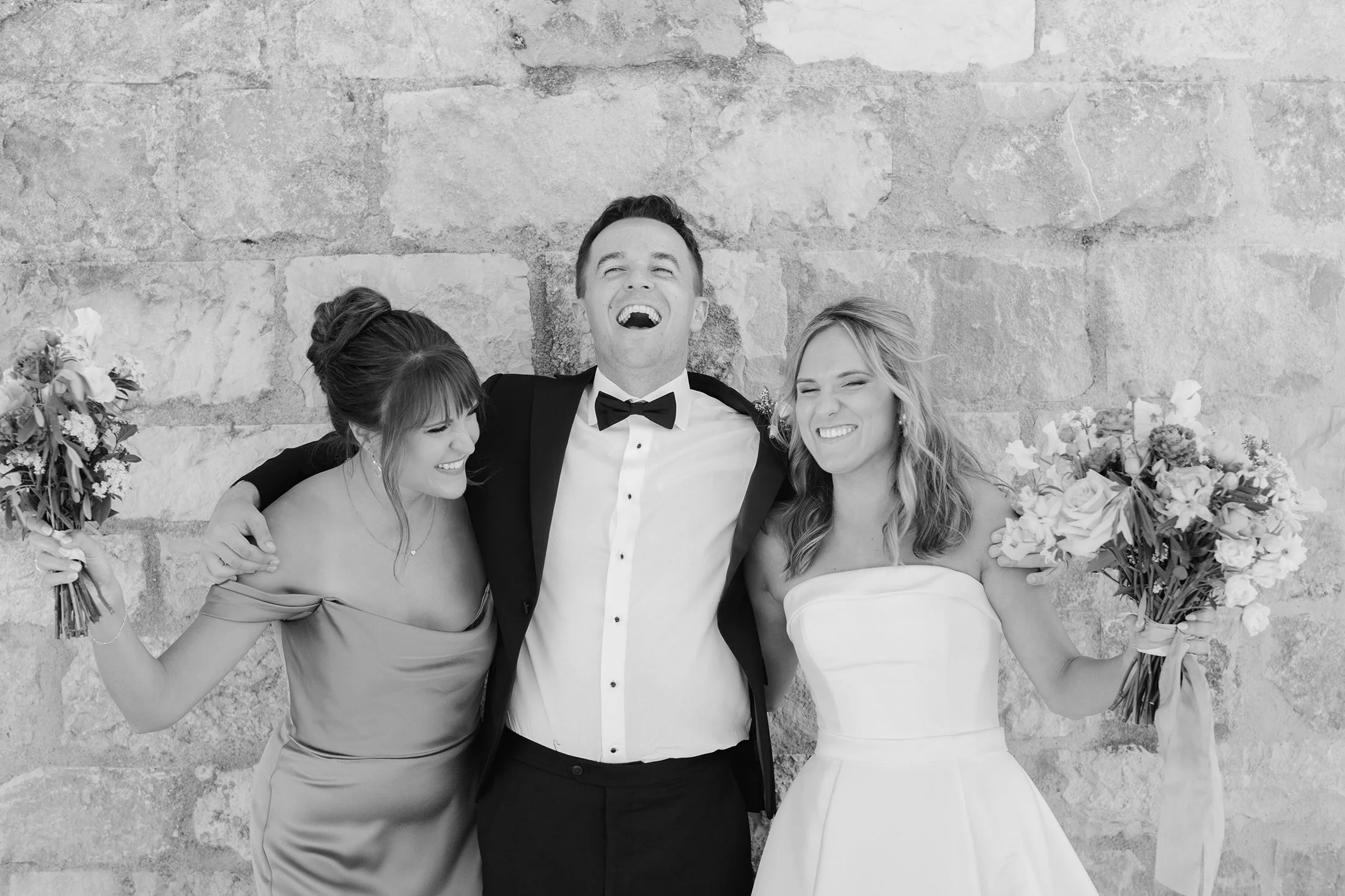 Black and white photo of groom and two bridesmaids laughing against a stone wall.