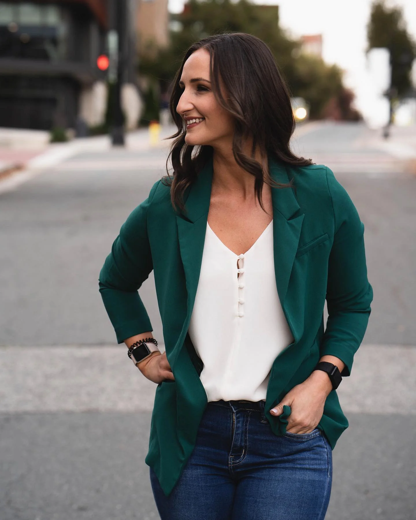 A woman with dark, wavy hair smiling and looking to her left, standing outdoors on a city street during daytime. She is wearing a green blazer, white blouse, dark jeans, and a smartwatch.