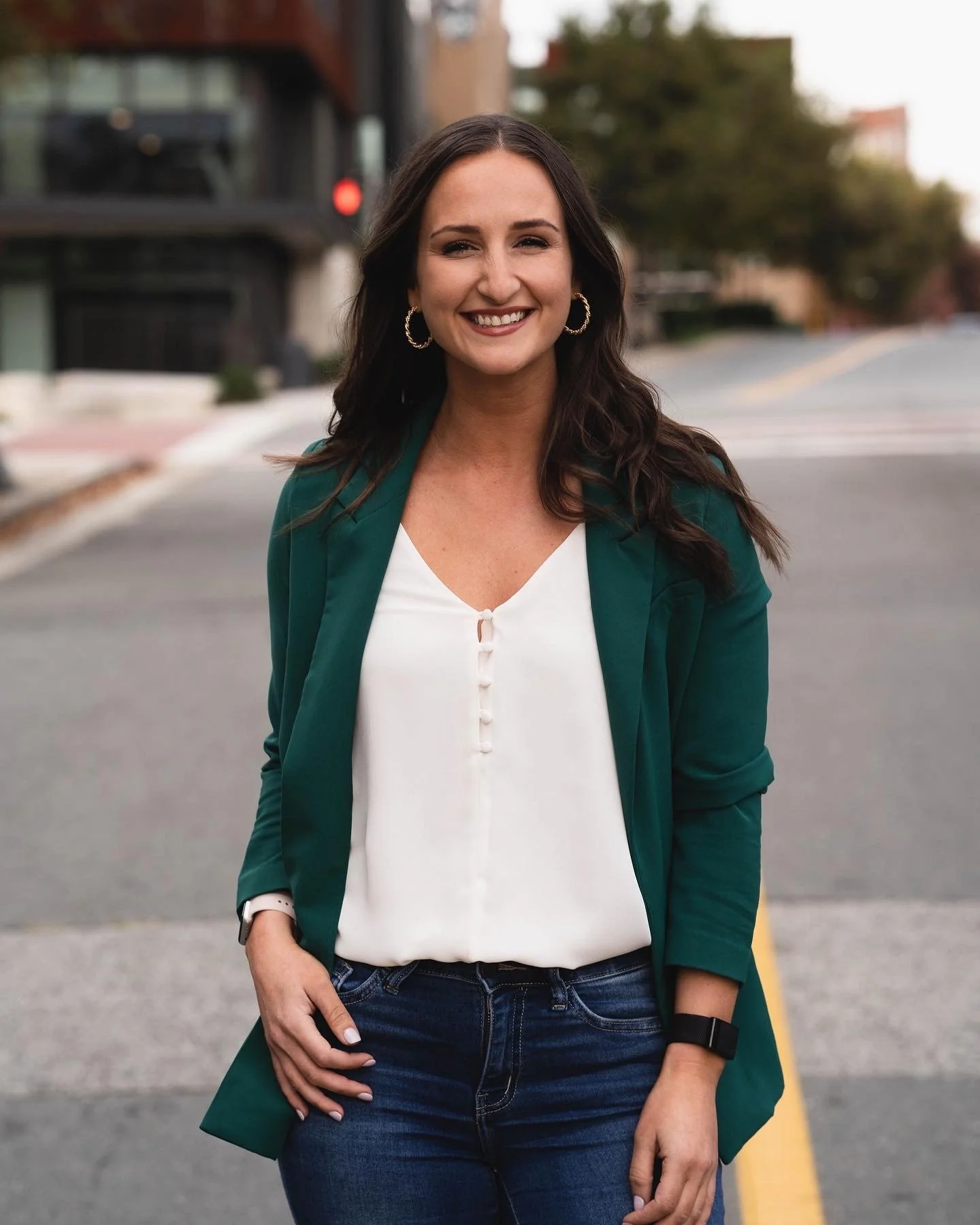 A woman smiling outdoors on a city street, wearing a green blazer, white blouse, and jeans, with dark wavy hair and hoop earrings.