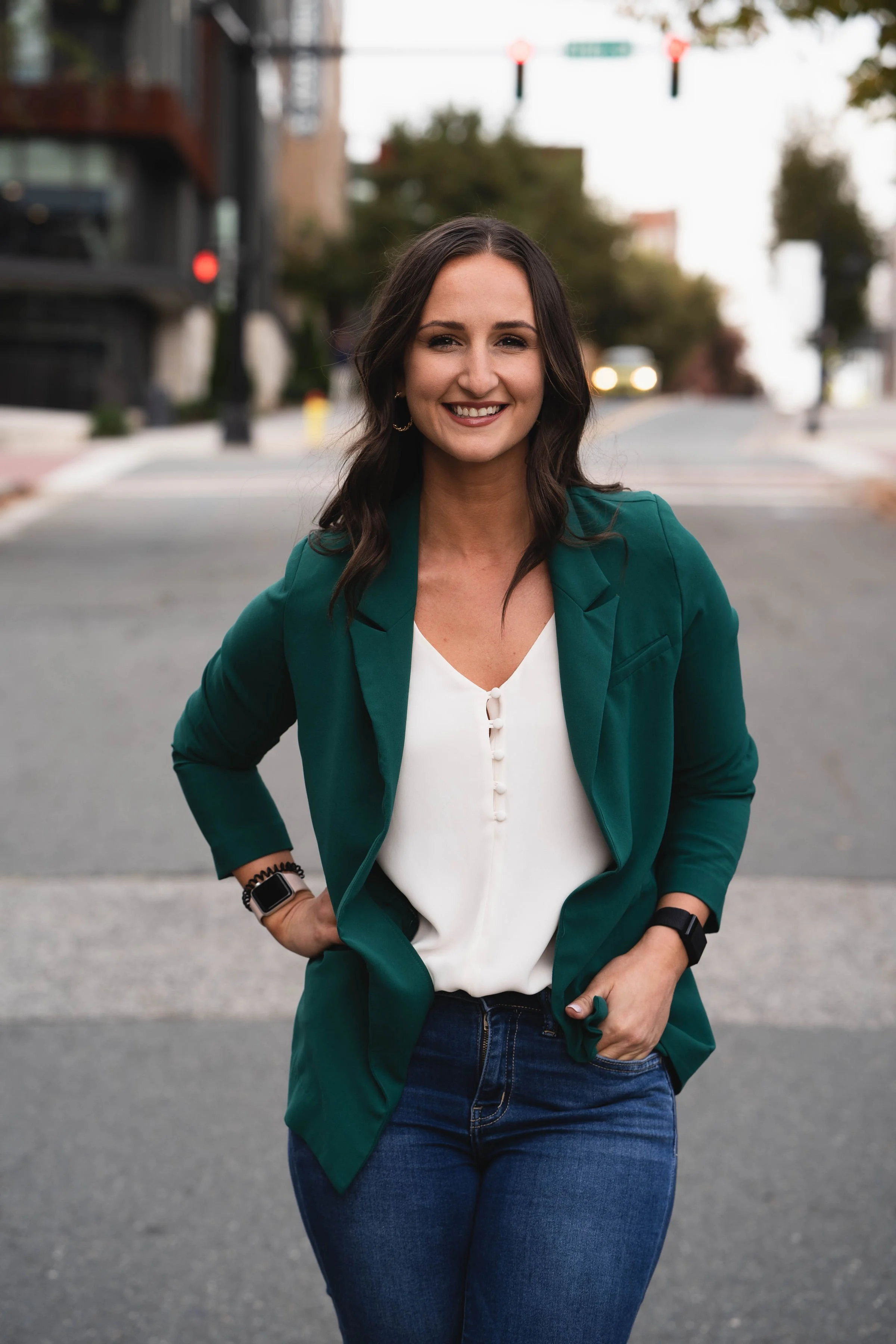 Smiling woman with dark hair, wearing a green blazer over a white blouse and blue jeans, standing on a city street with trees and traffic lights in the background.