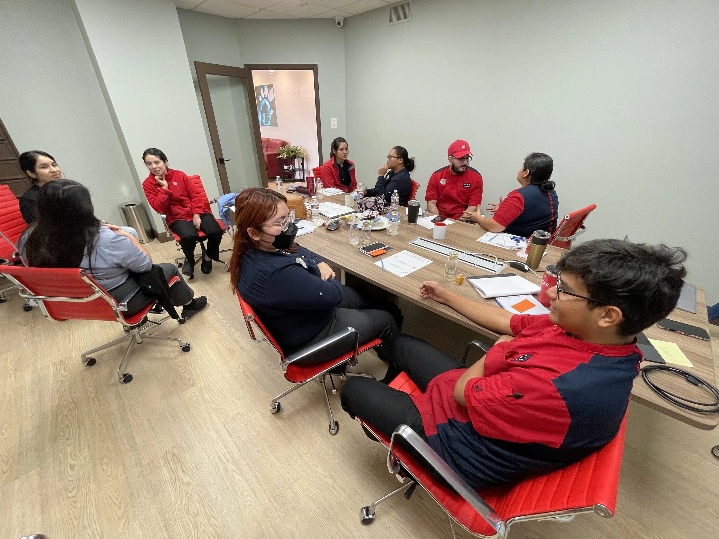 Group of people sitting around a large conference table in an office, engaging in discussion. Some are wearing red shirts, and there are papers, water bottles, and notebooks on the table.