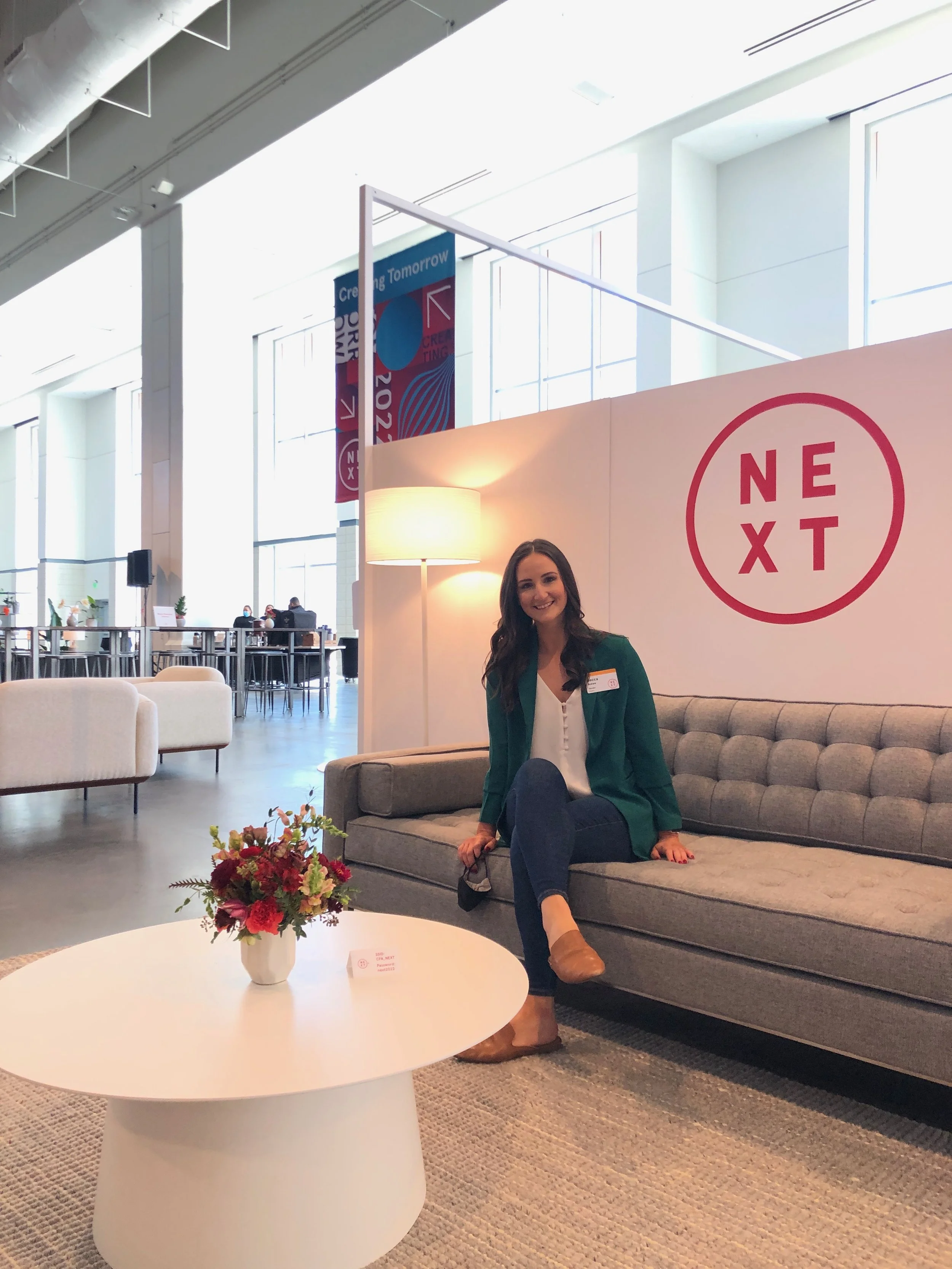 A woman with long dark hair, wearing a green blazer and brown boots, sits on a gray couch in a modern, brightly lit airport lounge. She is smiling at the camera, with a bouquet of flowers and a small sign on a white round table in front of her. The lounge has white walls with a large sign reading 'NEXT', high windows, and banners hanging from the ceiling.