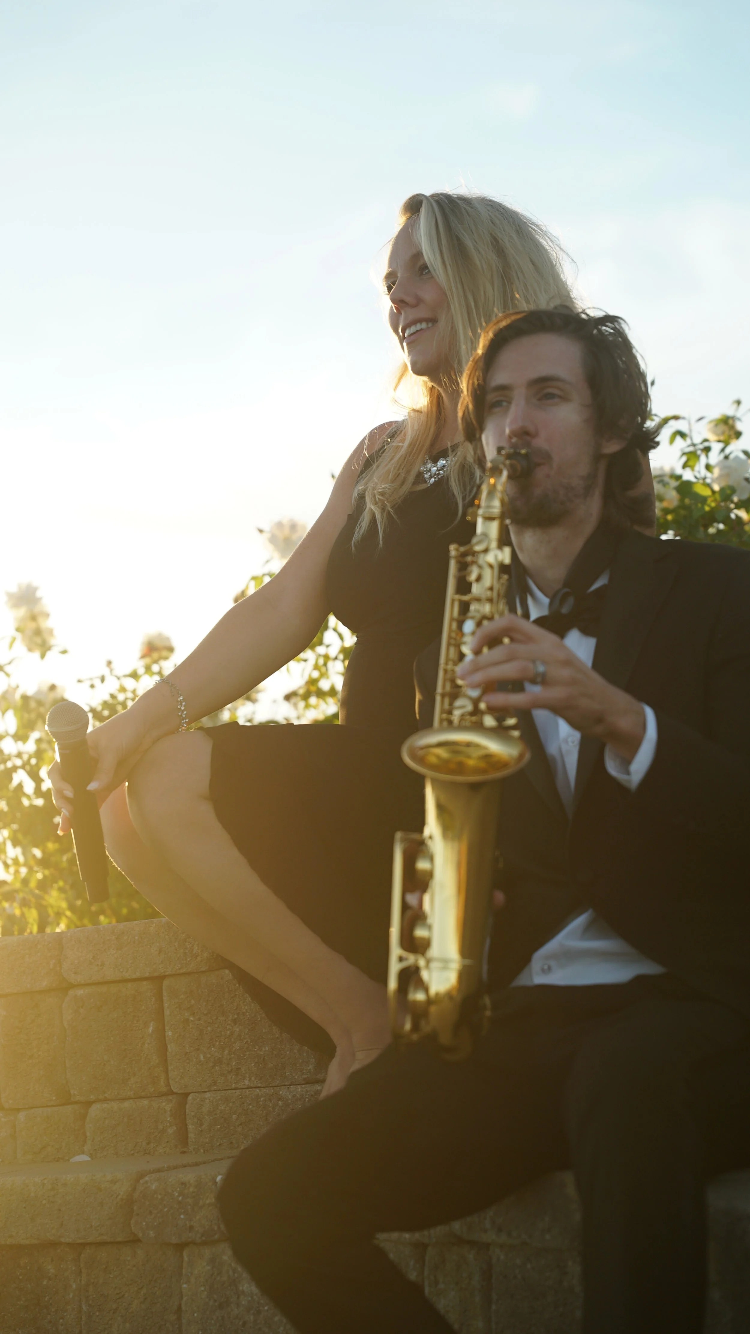 A woman and a man dressed formally, with the woman holding a microphone and the man playing a saxophone outdoors during sunset, with flowers in the background.
