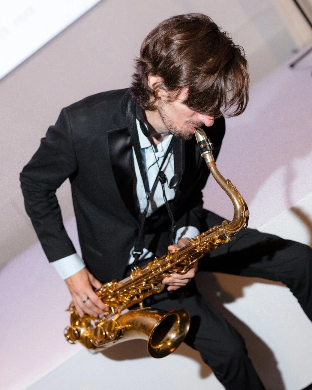 A man with long brown hair and a beard playing a gold-colored saxophone while seated on a light-colored surface.