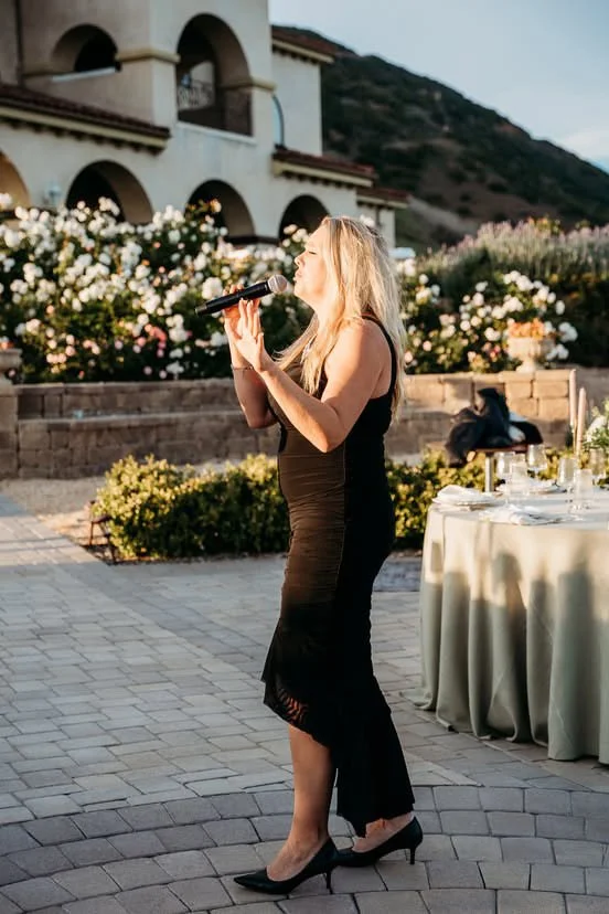 A woman in a black dress and heels sings into a microphone outdoors during sunset, with a backdrop of flowers, a building with arches, and a table set for an event.