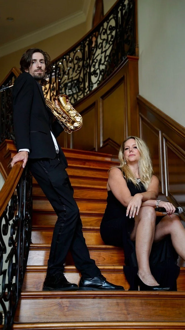 A man in a black suit holding a gold saxophone stands on a wooden staircase next to a woman in a black dress sitting on the stairs, with her arms crossed and a microphone in her hand.