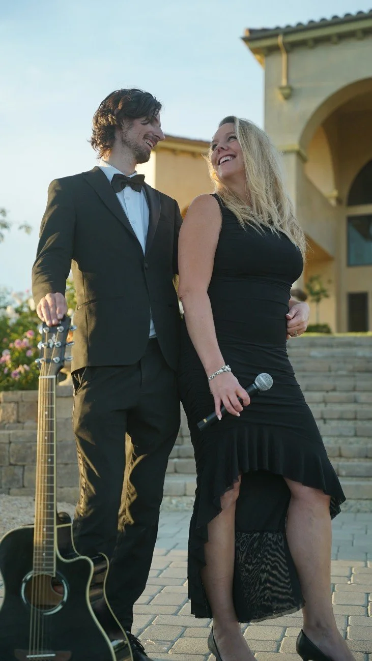 A man in a tuxedo and a woman in a black dress are smiling and looking at each other outdoors, with a guitar on the ground nearby and a building in the background.