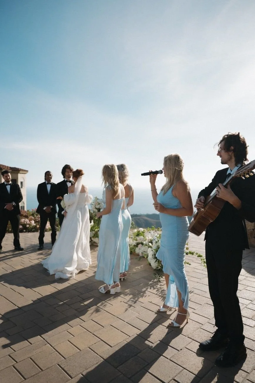 A wedding ceremony taking place outdoors on a sunny day with the bride and groom standing in front of a small group of bridesmaids and groomsmen, while a woman sings into a microphone and a man plays guitar. The scene is set on a brick patio with scenic views in the background.