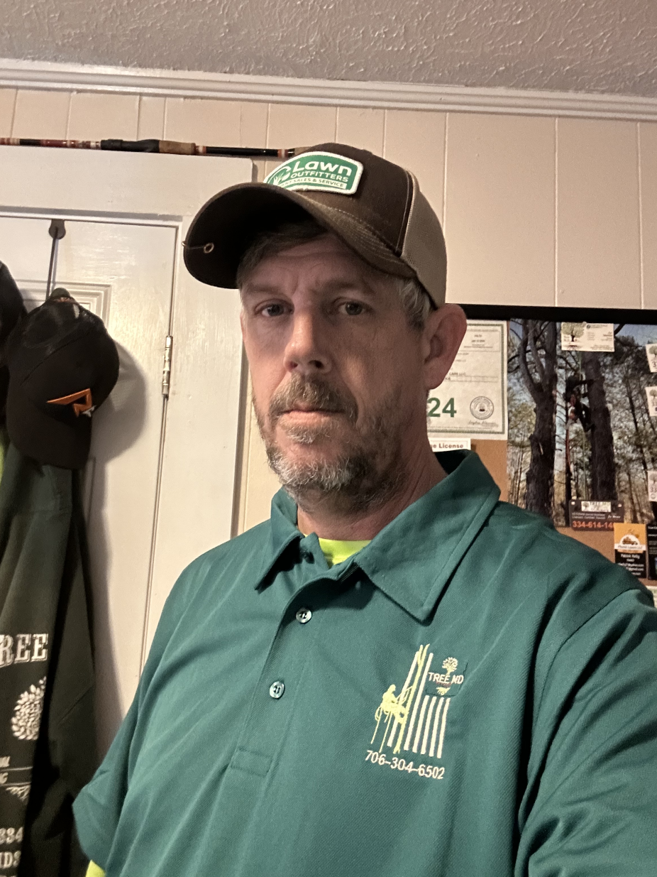 A man wearing a green polo shirt with a logo and phone number, a brown cap with a green and white logo, standing indoors near a white door with hooks and hanging items, behind him a corkboard with documents and a TV screen displaying trees.