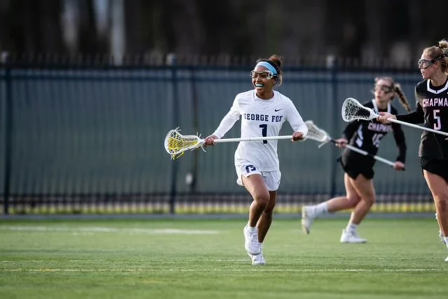 Coach Marena Tharpe competing in a white uniform with blue accents running on a field, holding a lacrosse stick, smiling, with three other players in black uniforms nearby.
