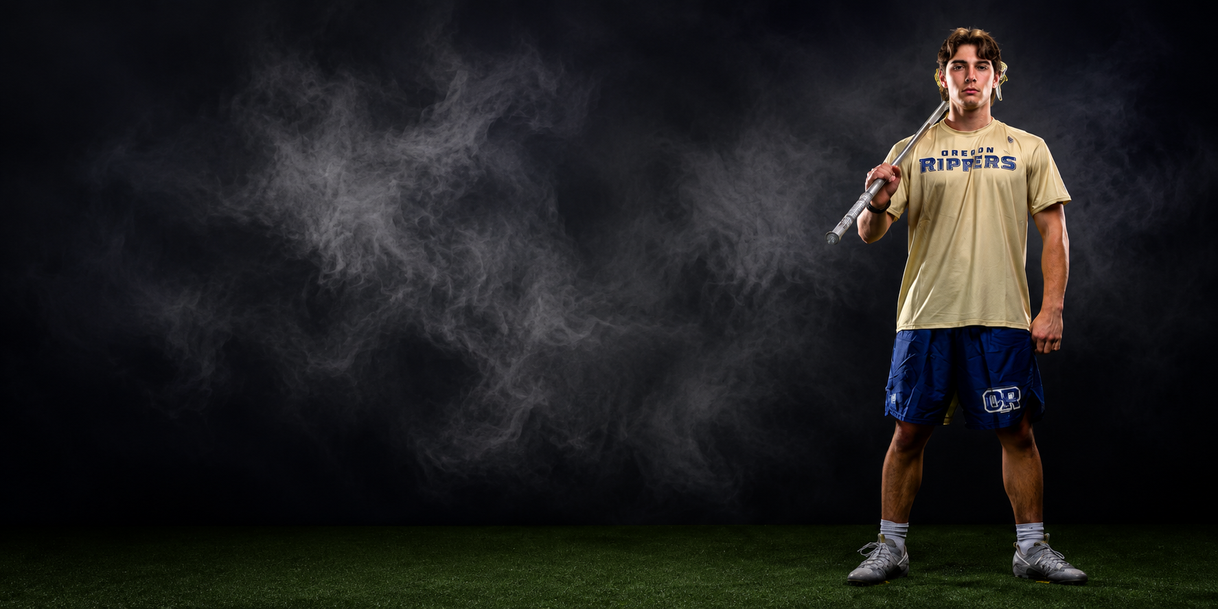 A young male athlete standing on a sports field at night, holding a lacrosse stick over his shoulder, wearing a beige jersey and blue shorts with the Rippers logo, with a dark background and fog or smoke around him.