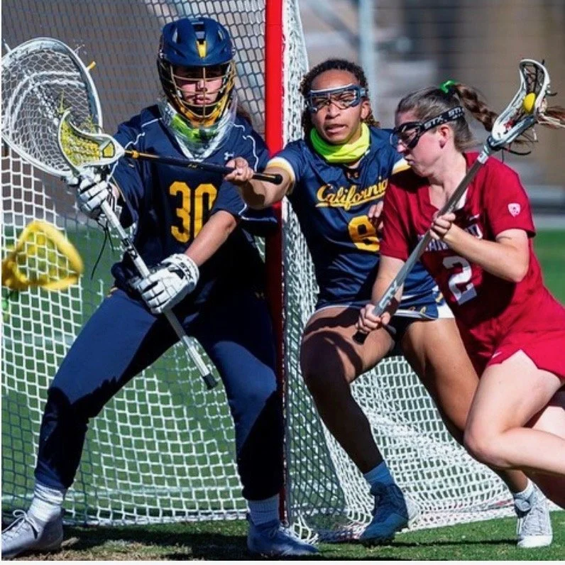 Coach Riley Drullinger competing near the goal, with one player in blue attempting to score, another in blue defending, and a player in red holding a lacrosse stick nearby.