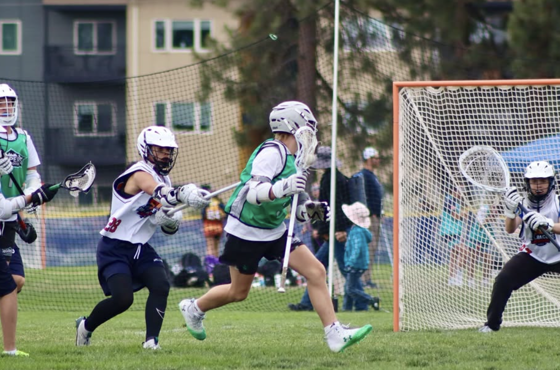 Rippers Oregon Lacrosse players in action on a grassy field during a game, with a goal and net in the background, and spectators watching.