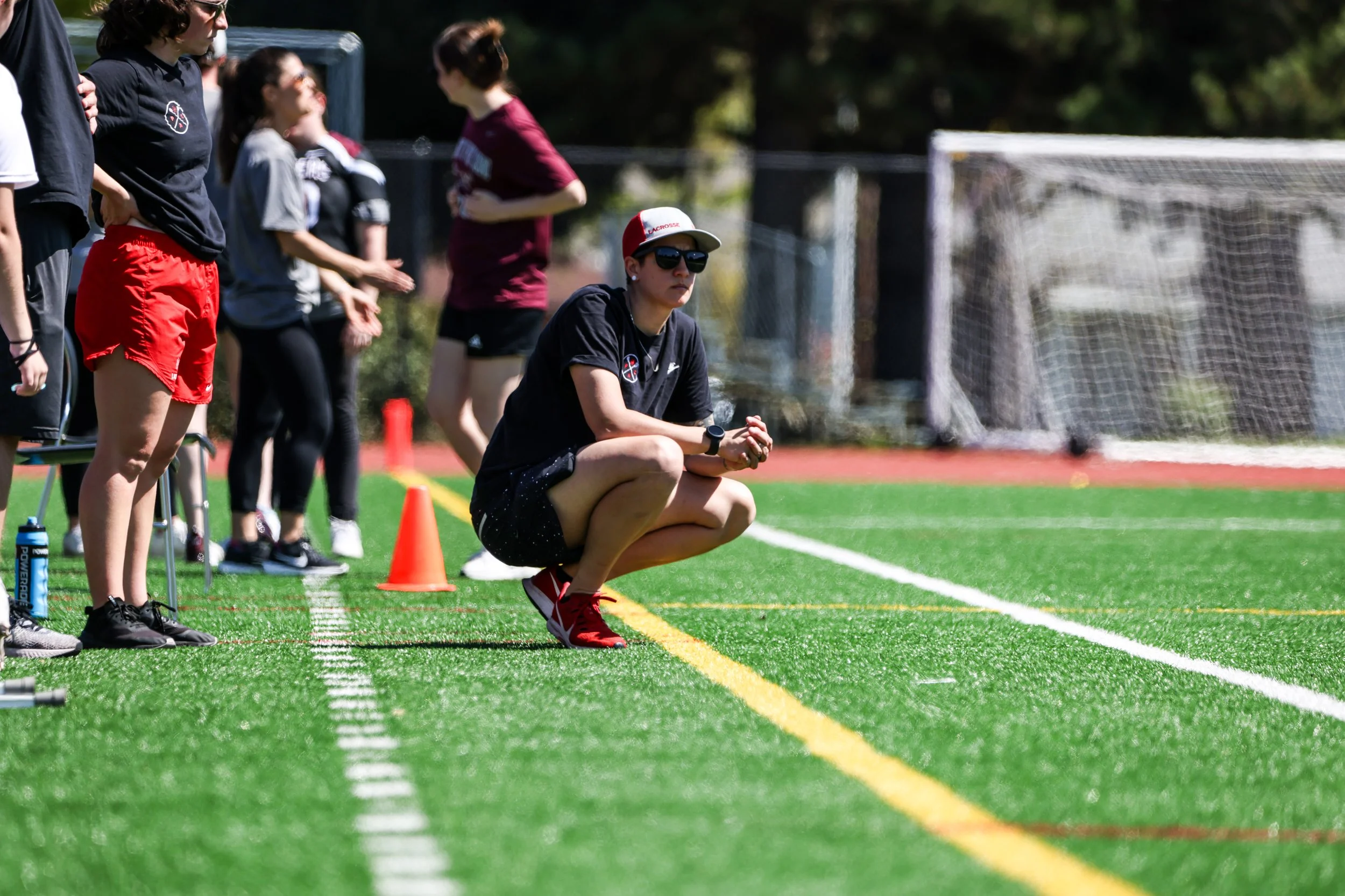 Coach Britt Hartman crouching on the sideline of a sports field, observing a game with players standing nearby. The scene is outdoors on a bright, sunny day.