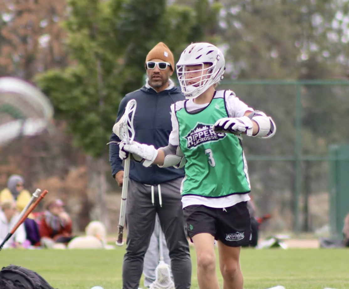 Rippers Oregon lacrosse player in a helmet, green jersey with the number 3, and shorts practicing on the field, while Coach Brian Silcott in the background.