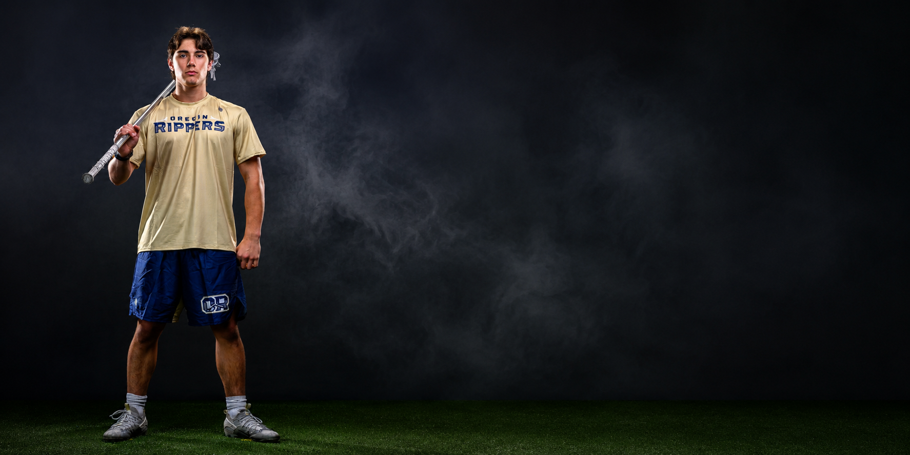 A young high school boy in sportswear holding a lacrosse stick over his shoulder, standing on a green turf against a dark background with smoke.