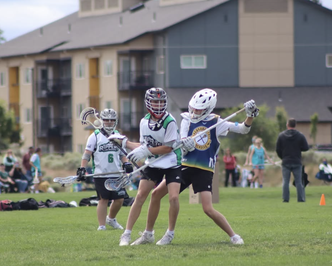 Rippers Oregon lacrosse players playing lacrosse on a grassy field, wearing helmets and sports gear, with spectators and apartment buildings in the background.